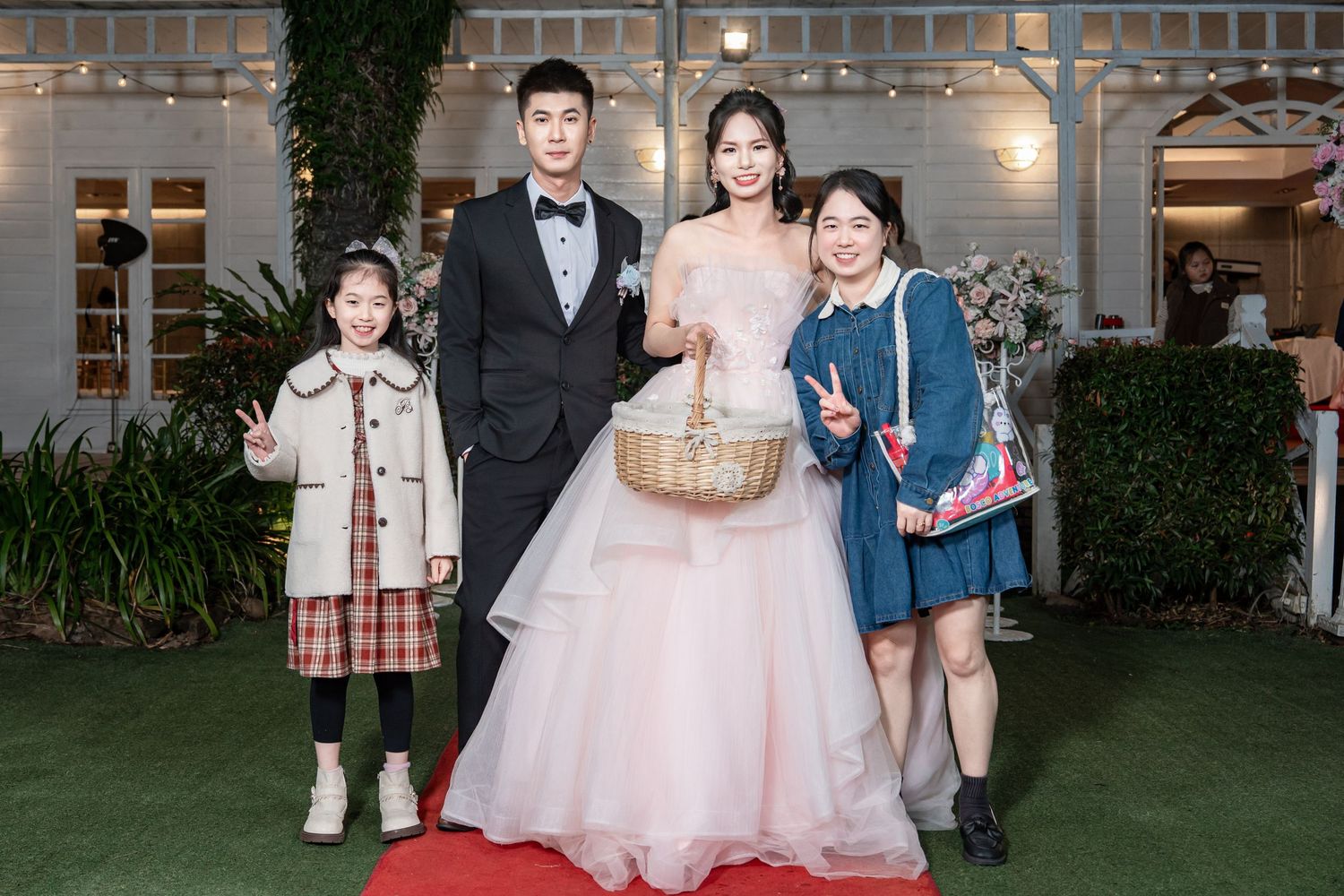 Group photo of wedding attendees gathered outside a venue with brick walls and decorative lighting.