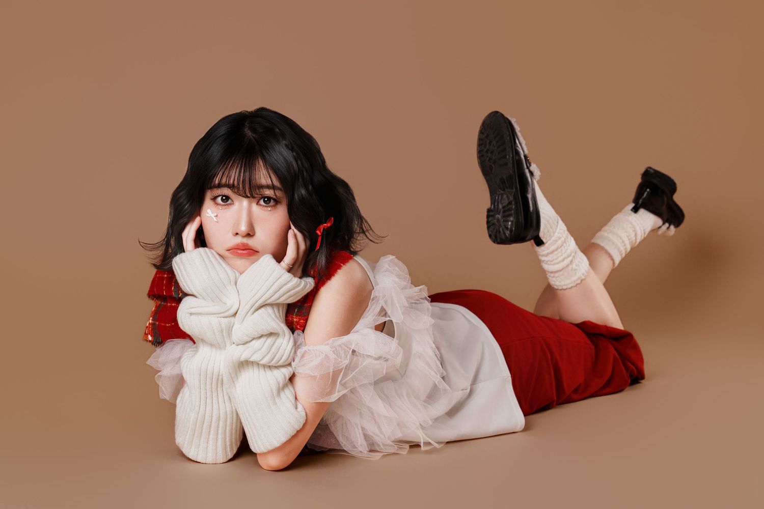 A person in a white sweater and red skirt lies on their side against a brown background, wearing black shoes and white socks.