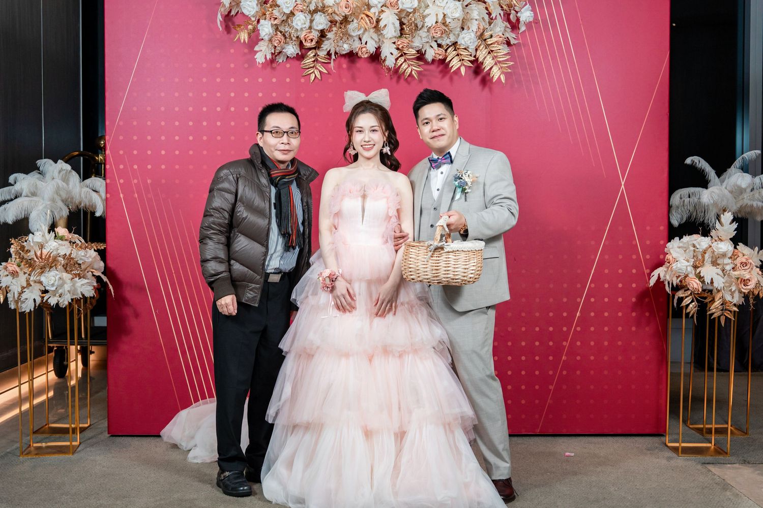 Wedding party poses in front of a pink backdrop with elegant floral chandelier decorations at a formal celebration.