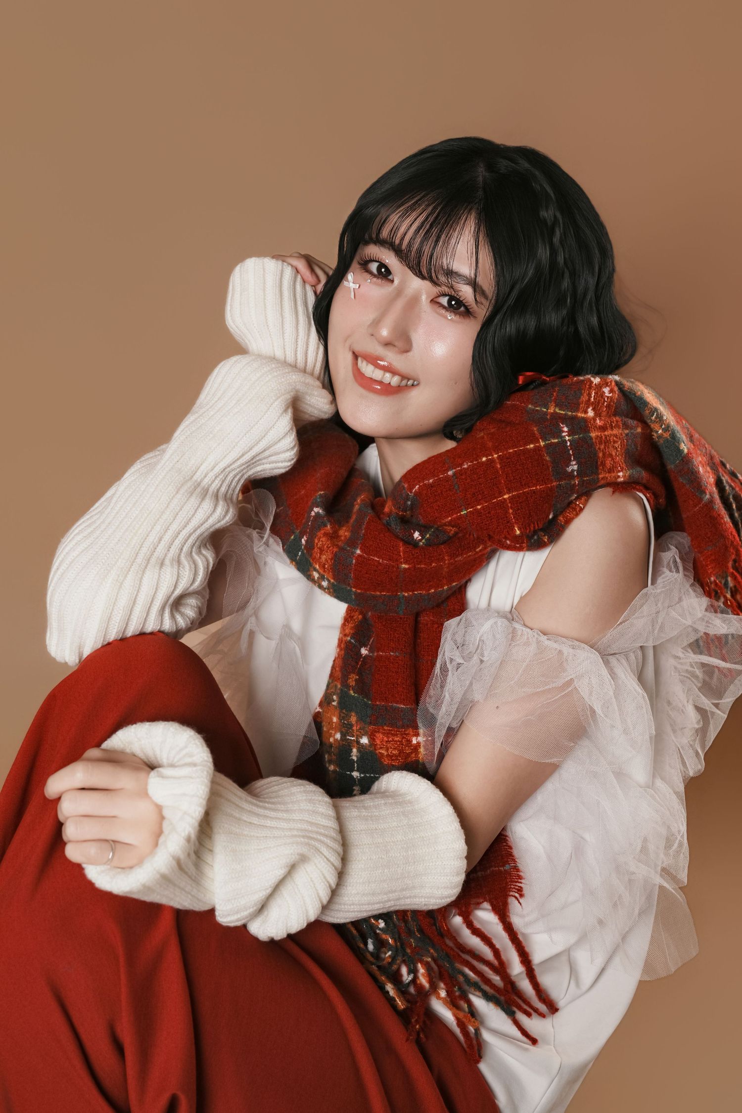 A person wearing a white sweater and red plaid scarf poses playfully against a brown background.