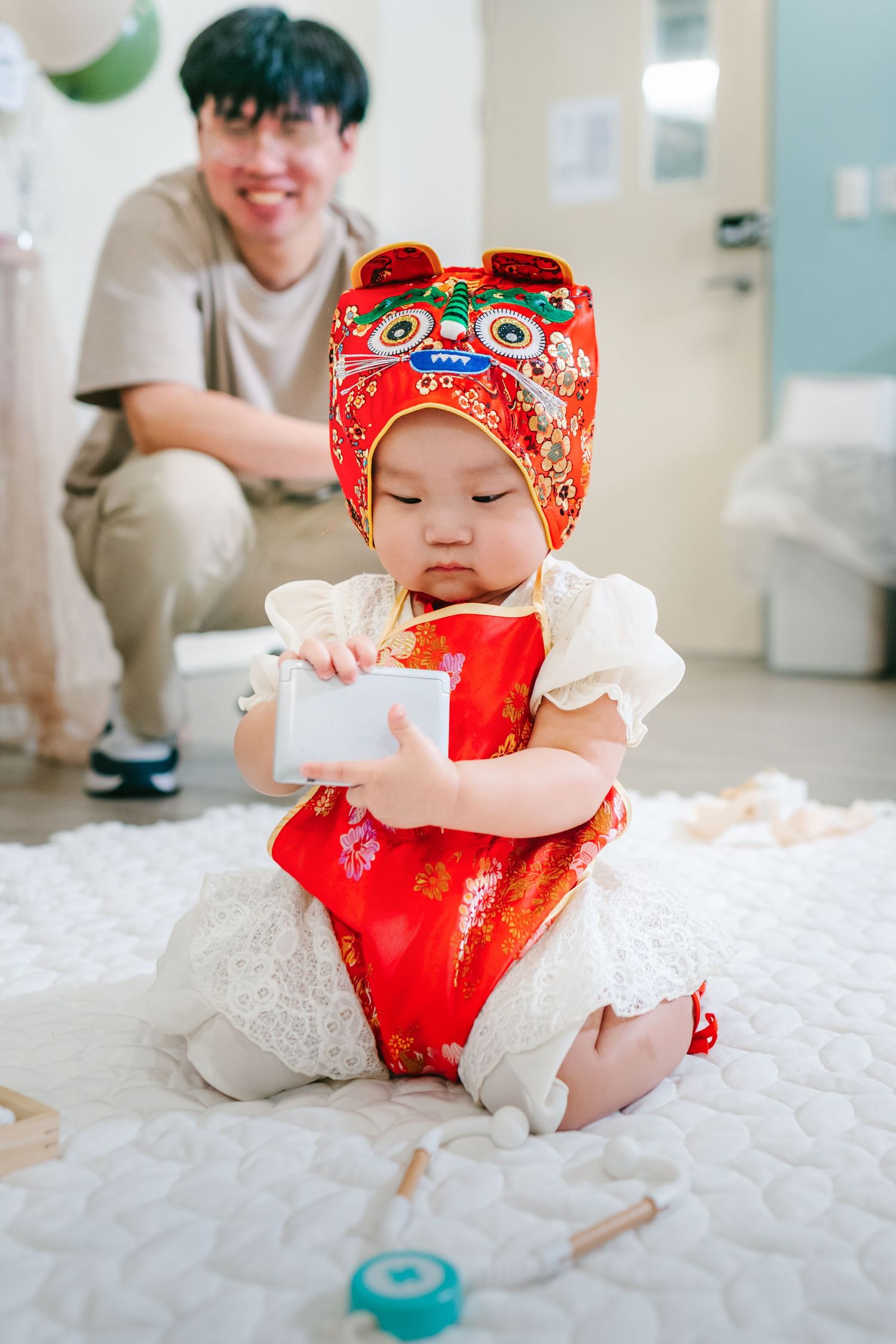 Baby wearing traditional red Chinese outfit and ornate cap sits on white carpet while holding a card.