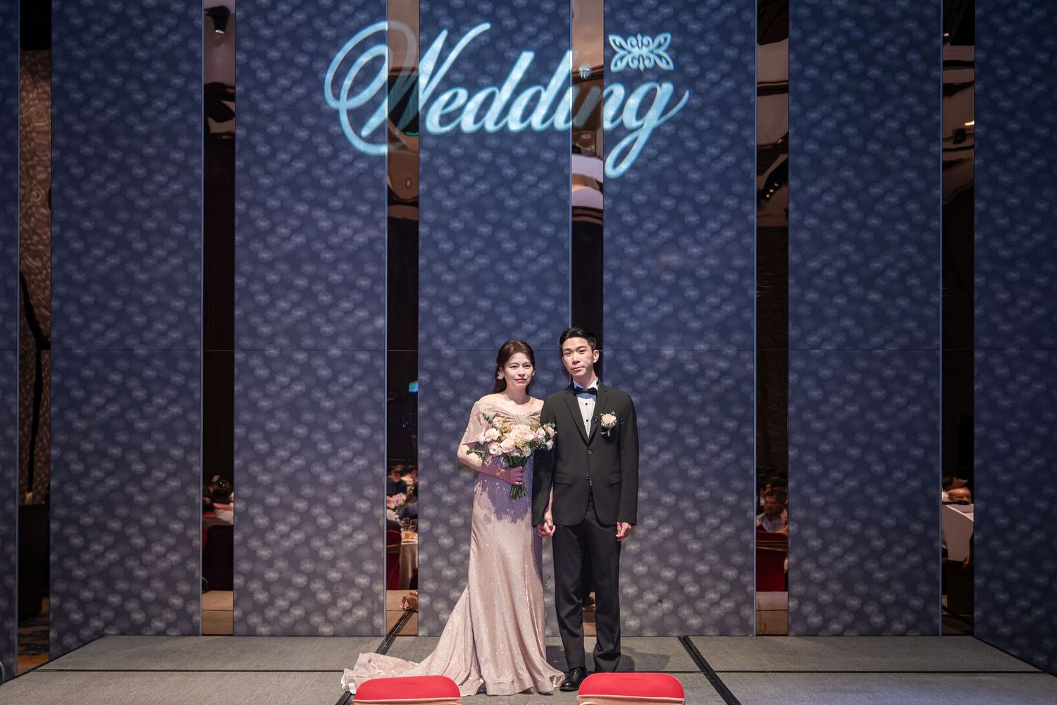 A couple stands on a red carpet stage with a glowing blue 'Wedding' sign illuminated behind them.