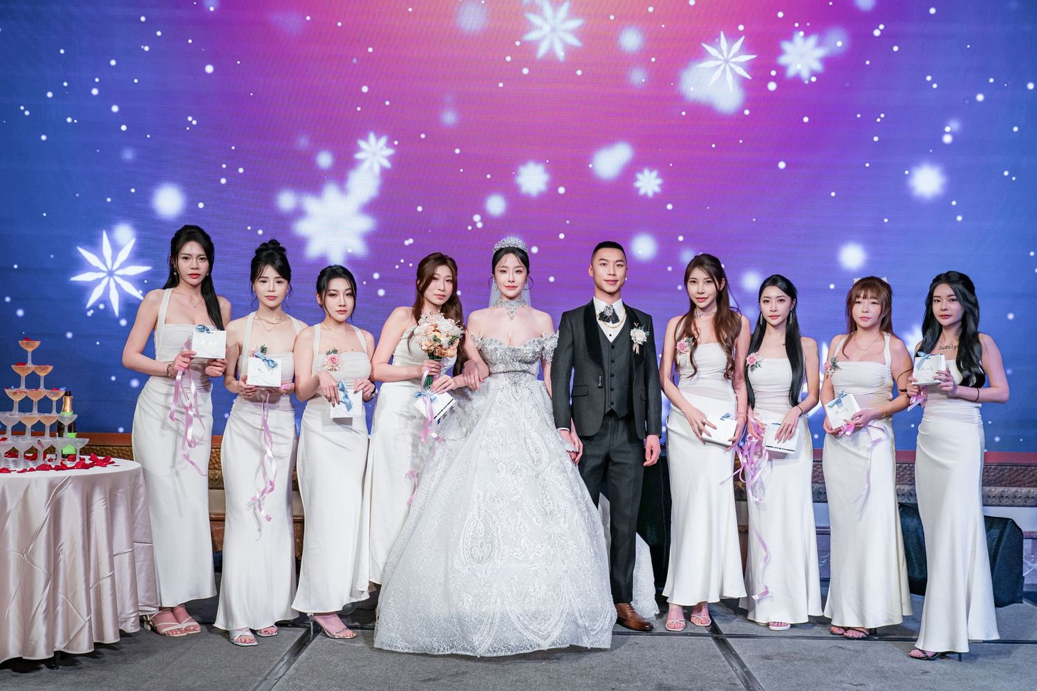 Wedding party members in white dresses and formal attire pose together against a blue and purple illuminated backdrop.