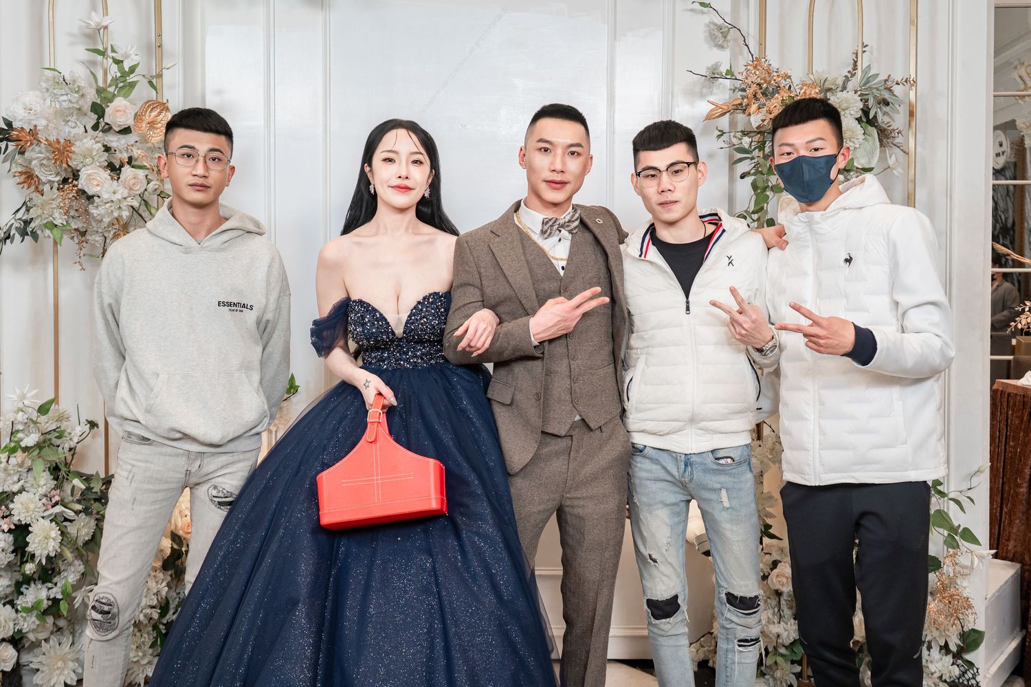 Group of wedding guests pose together in formal attire against floral backdrop at celebration event.