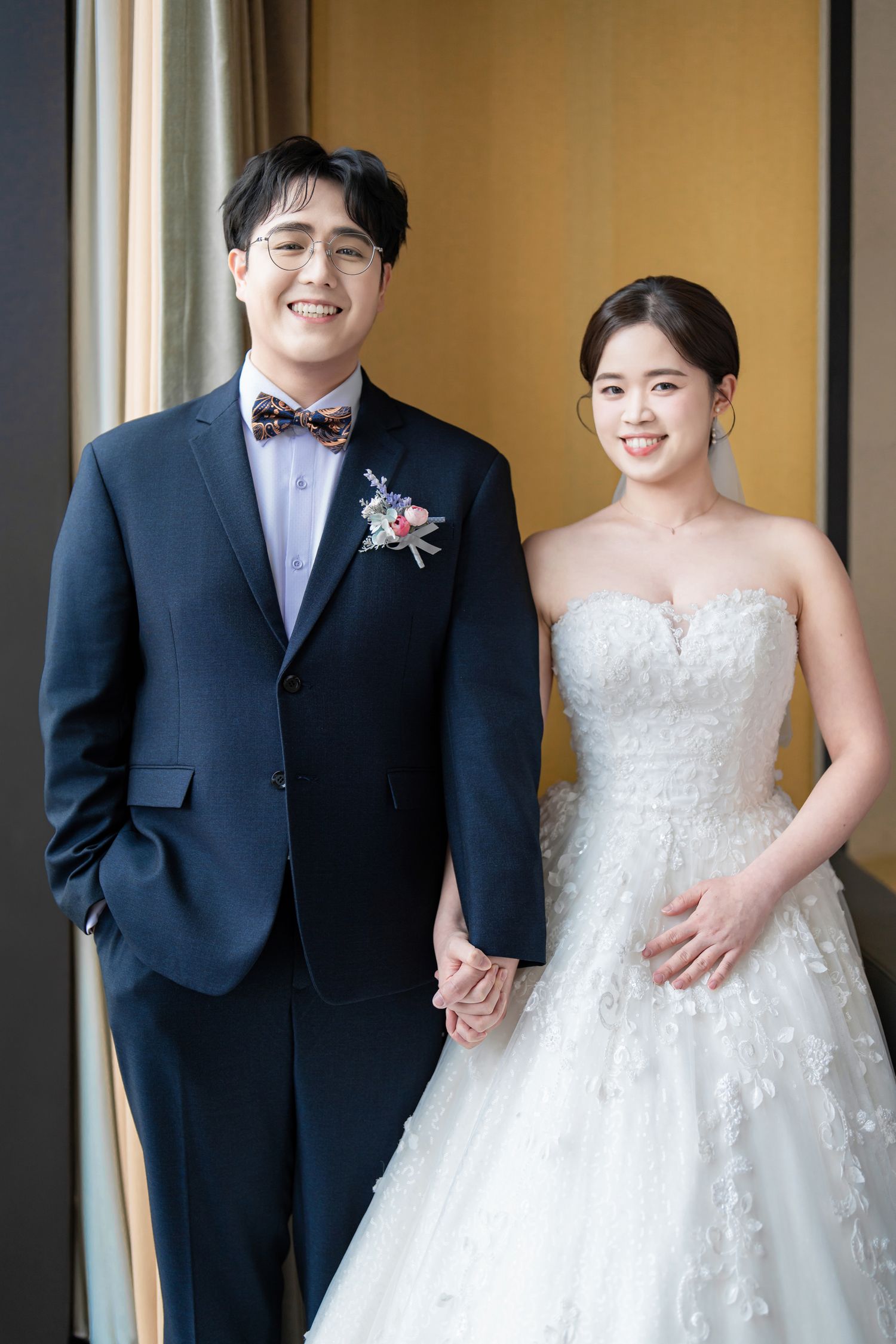 A couple poses together in wedding attire, with the bride in a white lace gown and groom in a dark suit.