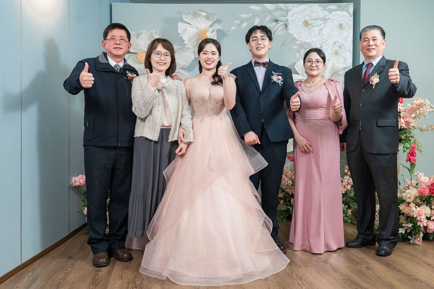 A group poses together in formal attire with pink dresses and dark suits against a blue wall with floral decorations.