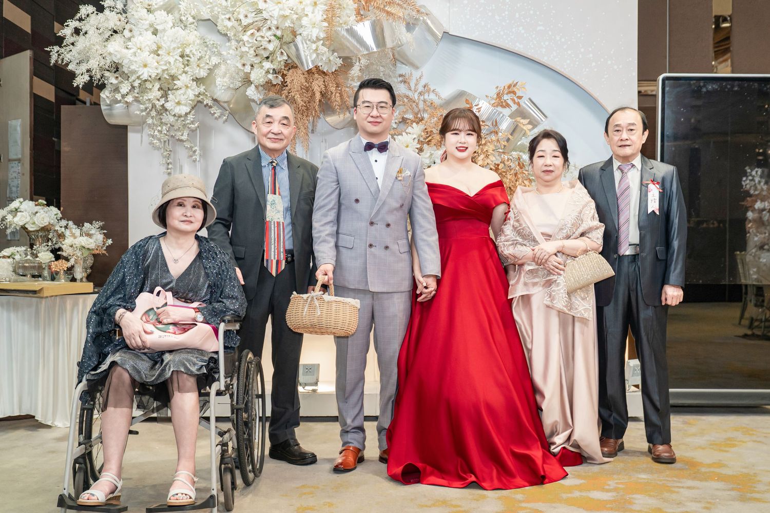 Wedding party members in formal attire pose together with red and neutral colored dresses in an elegant venue.