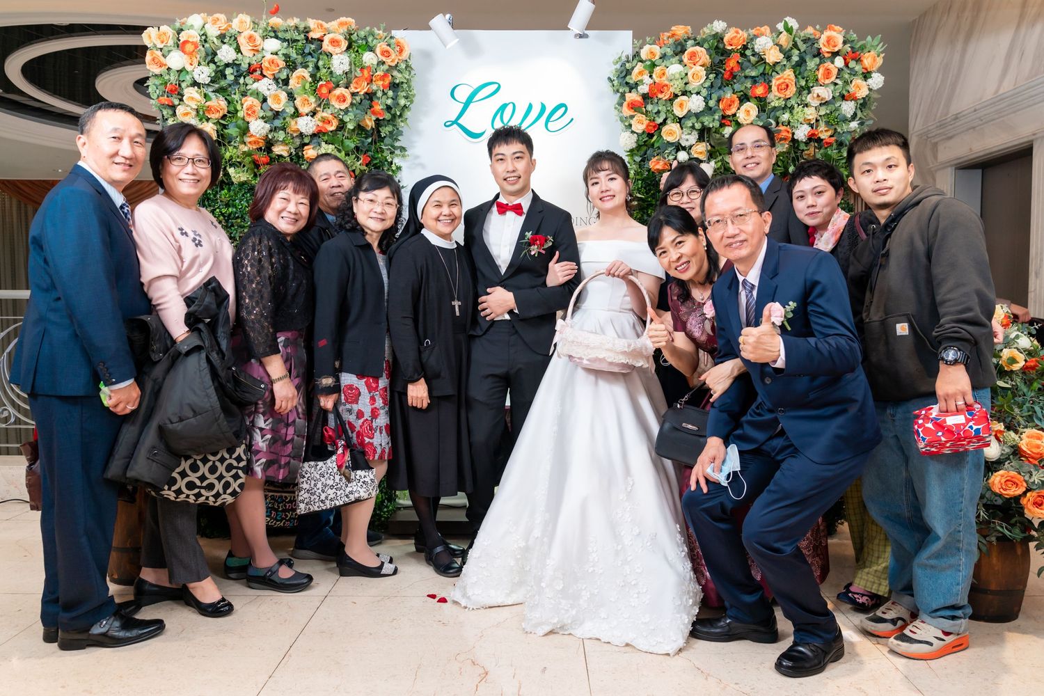 Large group wedding photo with guests gathered in front of floral wall decoration.