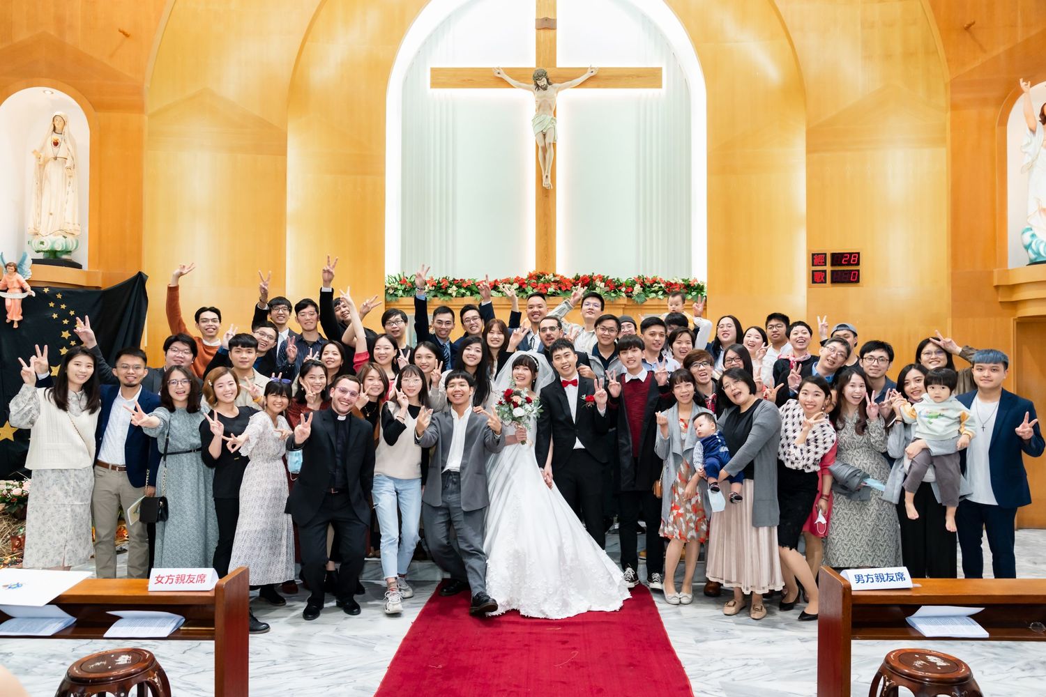 Festive wedding group photo with guests making peace signs in golden-walled church.