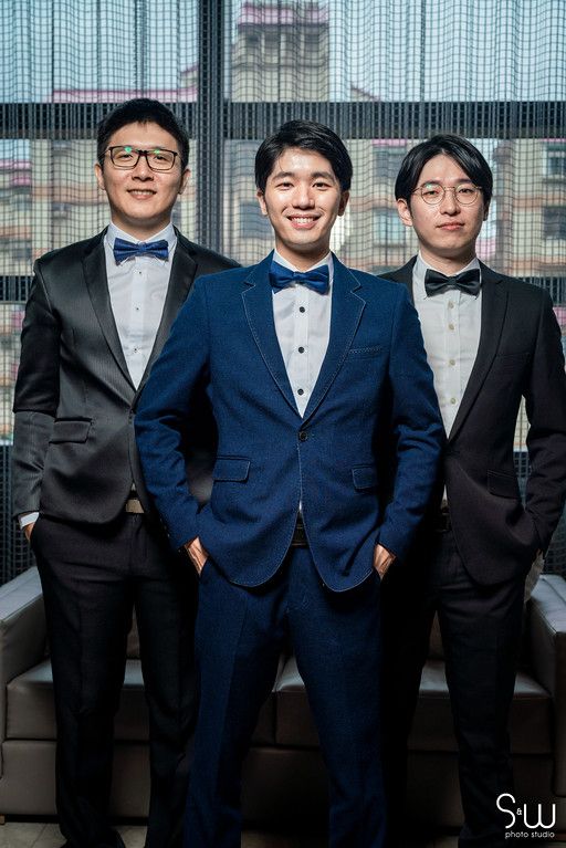 Three men in tuxedos pose formally by a window, with the center one wearing a navy blue suit.