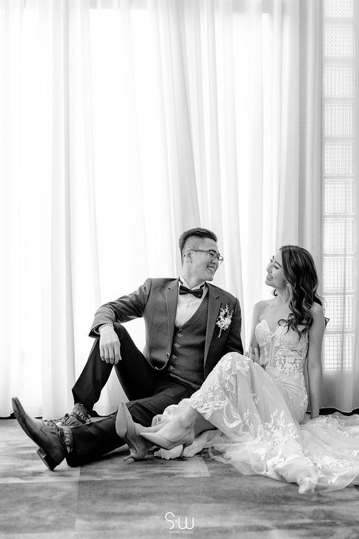 Black and white wedding photo showing couple sitting casually by bright window.