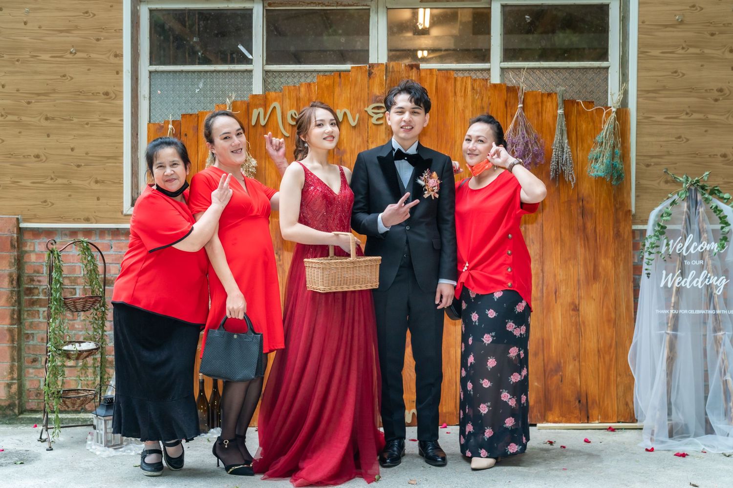 Group poses in front of wooden backdrop wearing formal attire with red accents and holding wicker basket.