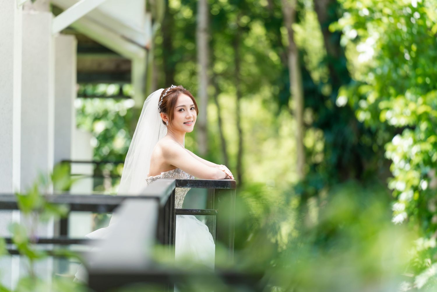 Bride in flowing white gown looks serene while resting on balcony railing.
