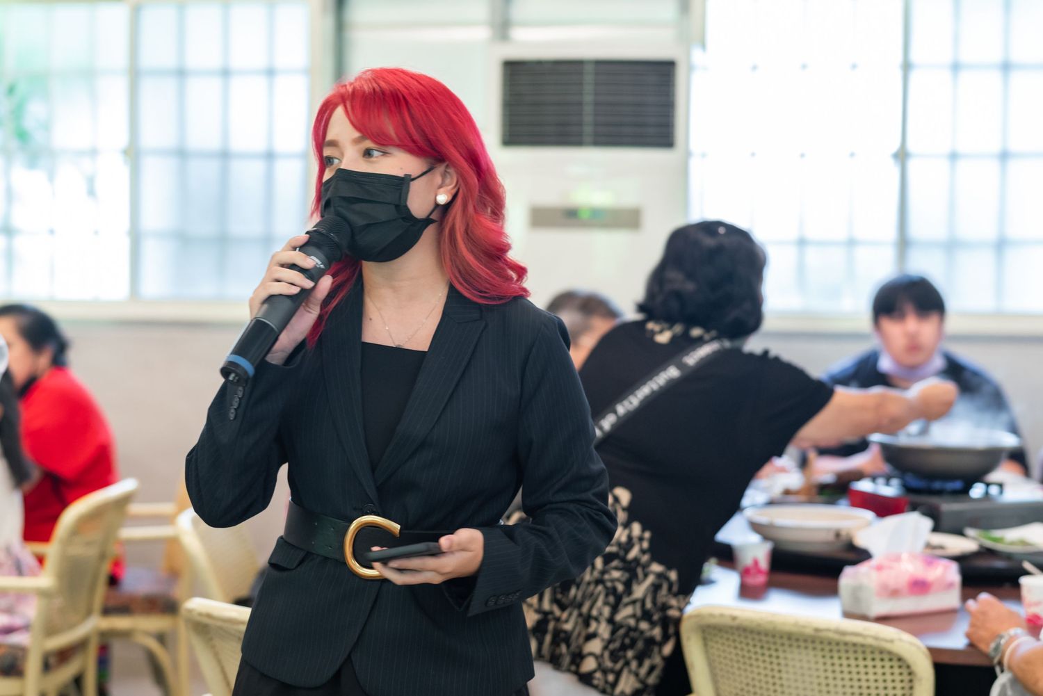 Speaker in black suit and face mask addressing audience at indoor event.
