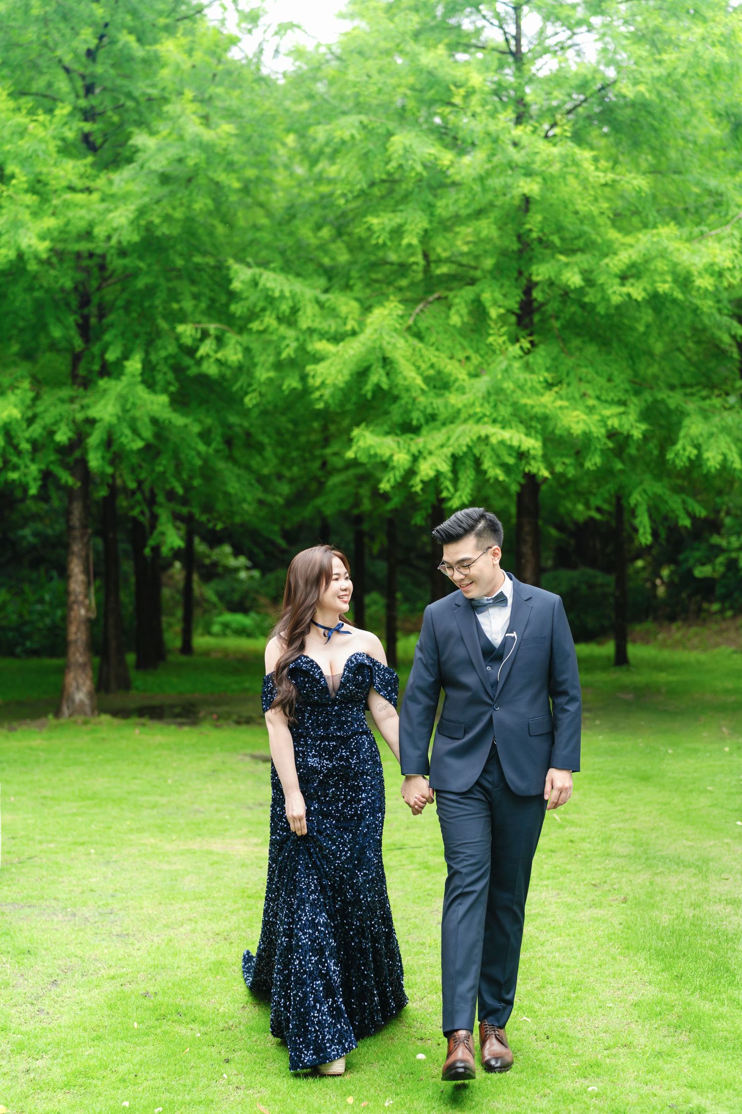 Couple walks hand in hand through a green summer park wearing formal attire - a blue gown and navy suit.