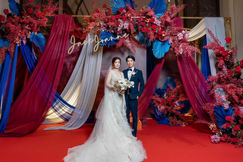Elegant wedding ceremony setup with red and blue floral arch, draped burgundy and white fabric, and a vibrant red carpet.