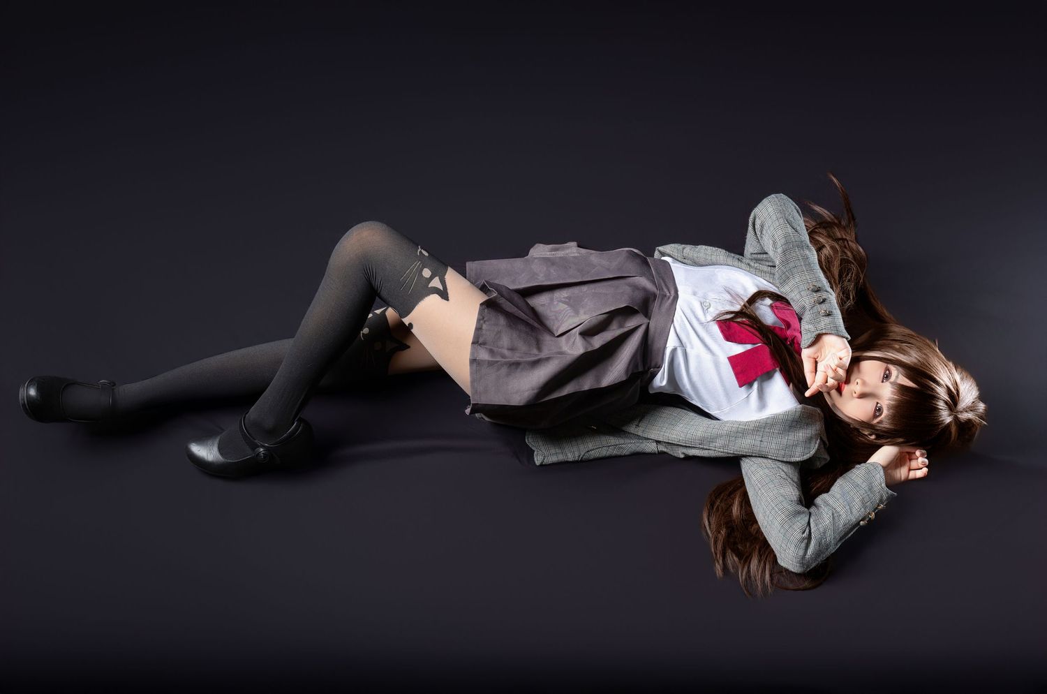 A model laying on dark floor wearing school uniform style clothing with long black stockings and red bow.