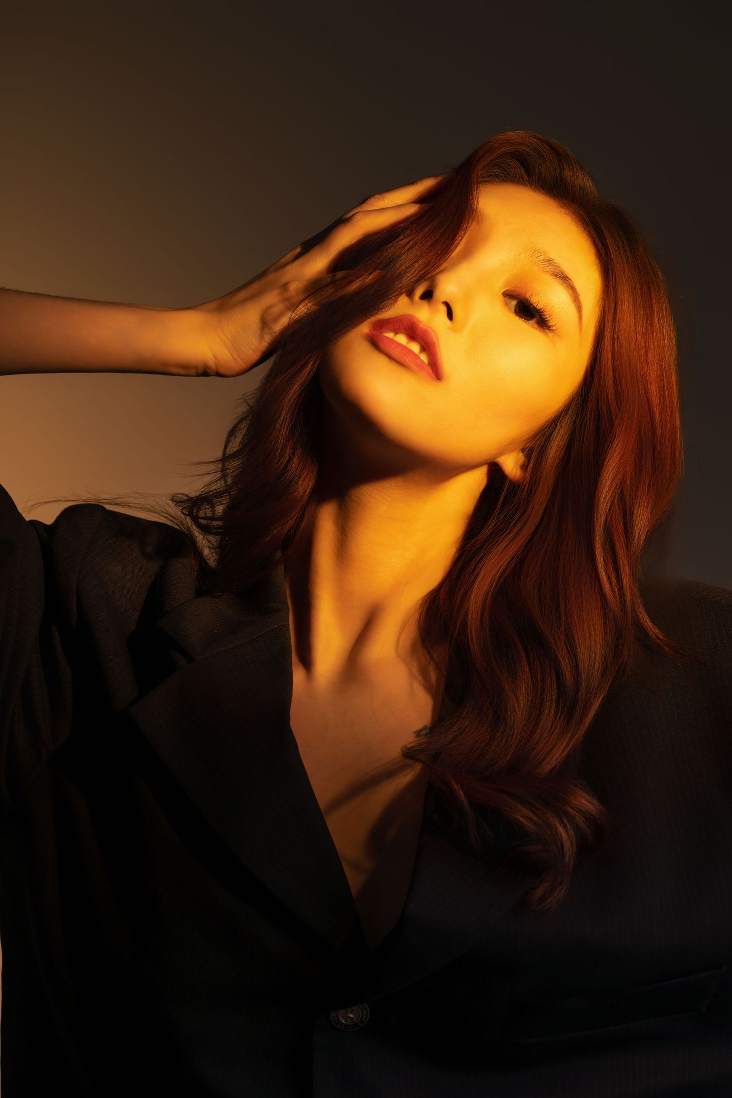 Portrait in moody warm lighting with dramatic shadows and black clothing against dark background.