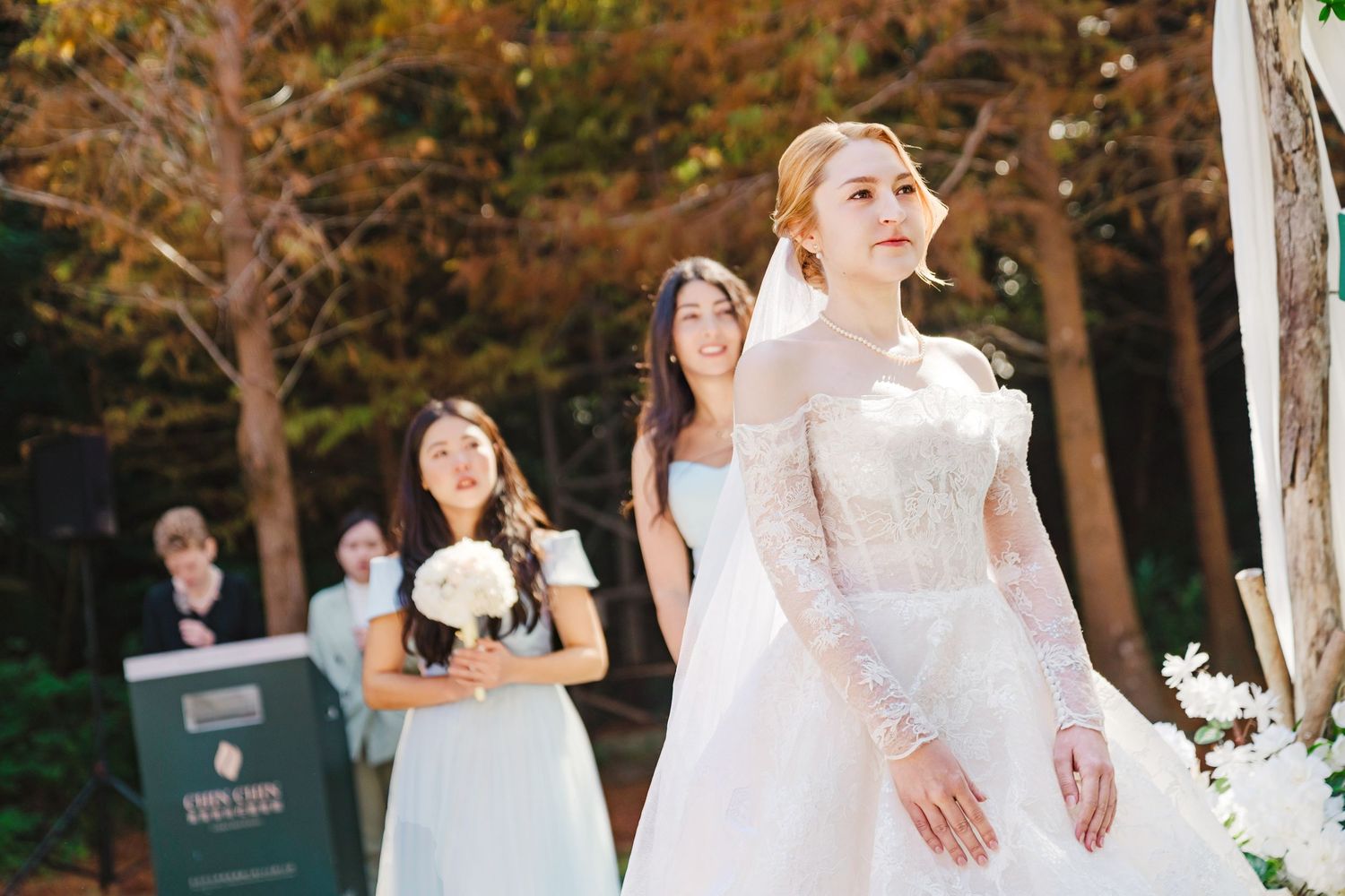 A bride in a long-sleeved lace wedding gown stands outdoors in an autumn forest setting during a ceremony.