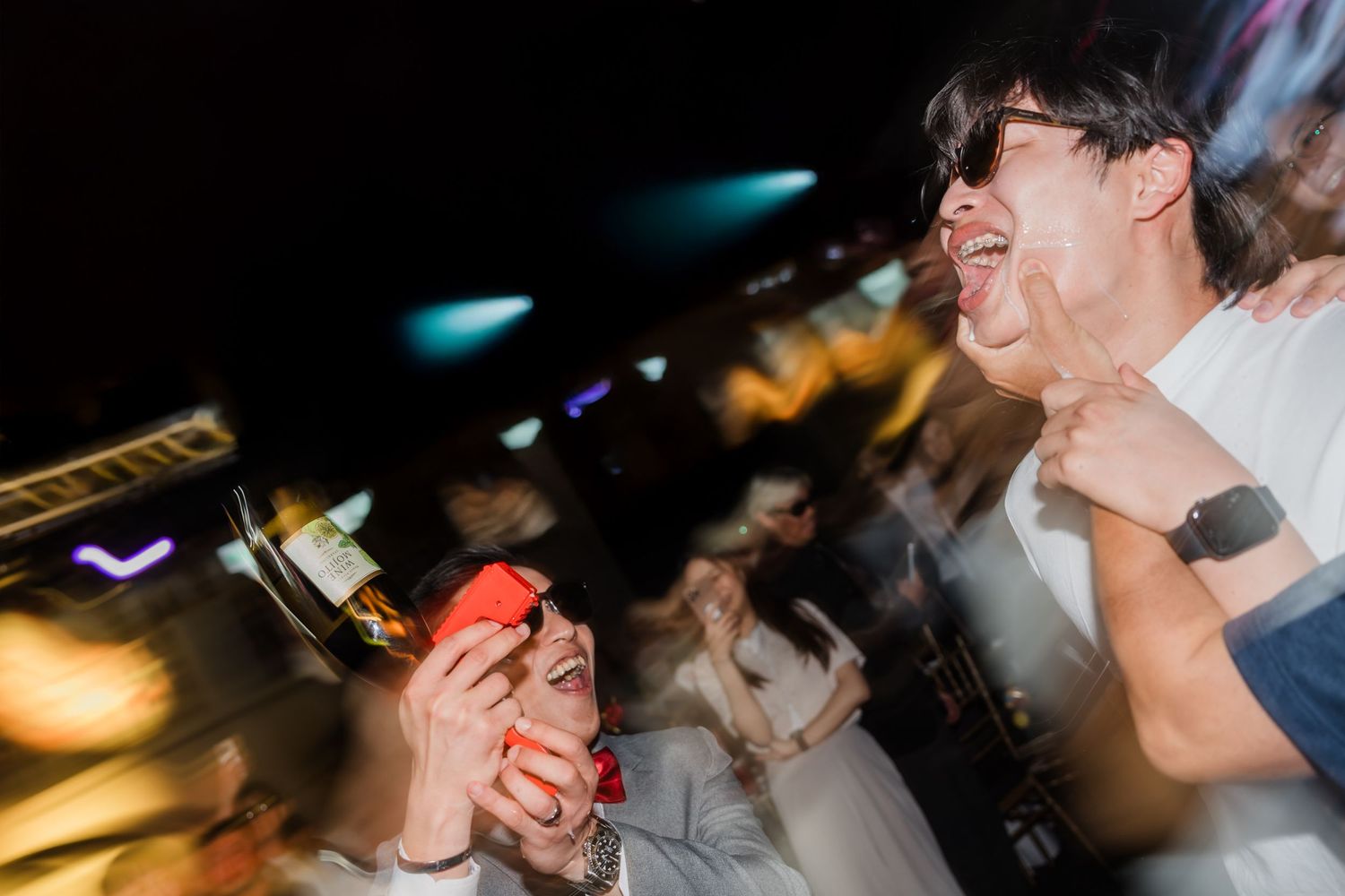 Motion-blurred photo of people dancing and celebrating at a nightclub with colorful lights in the background.