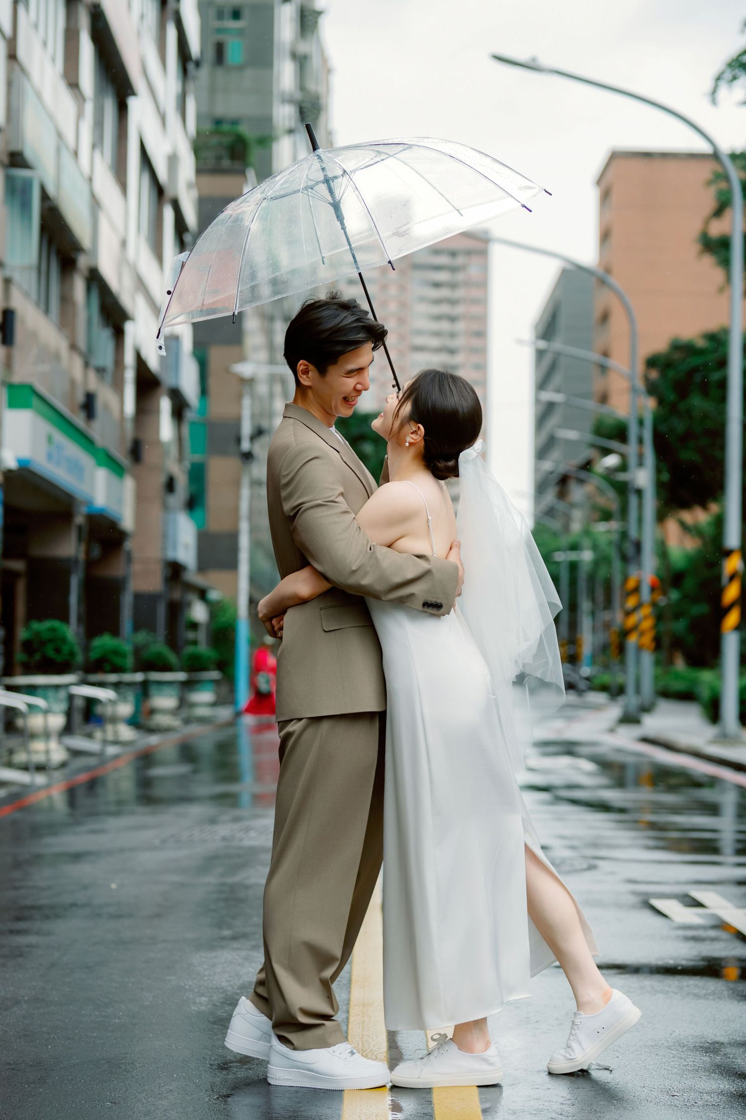 A romantic couple shares an umbrella on a rainy city street, the bride in white and groom in beige sharing an intimate moment.