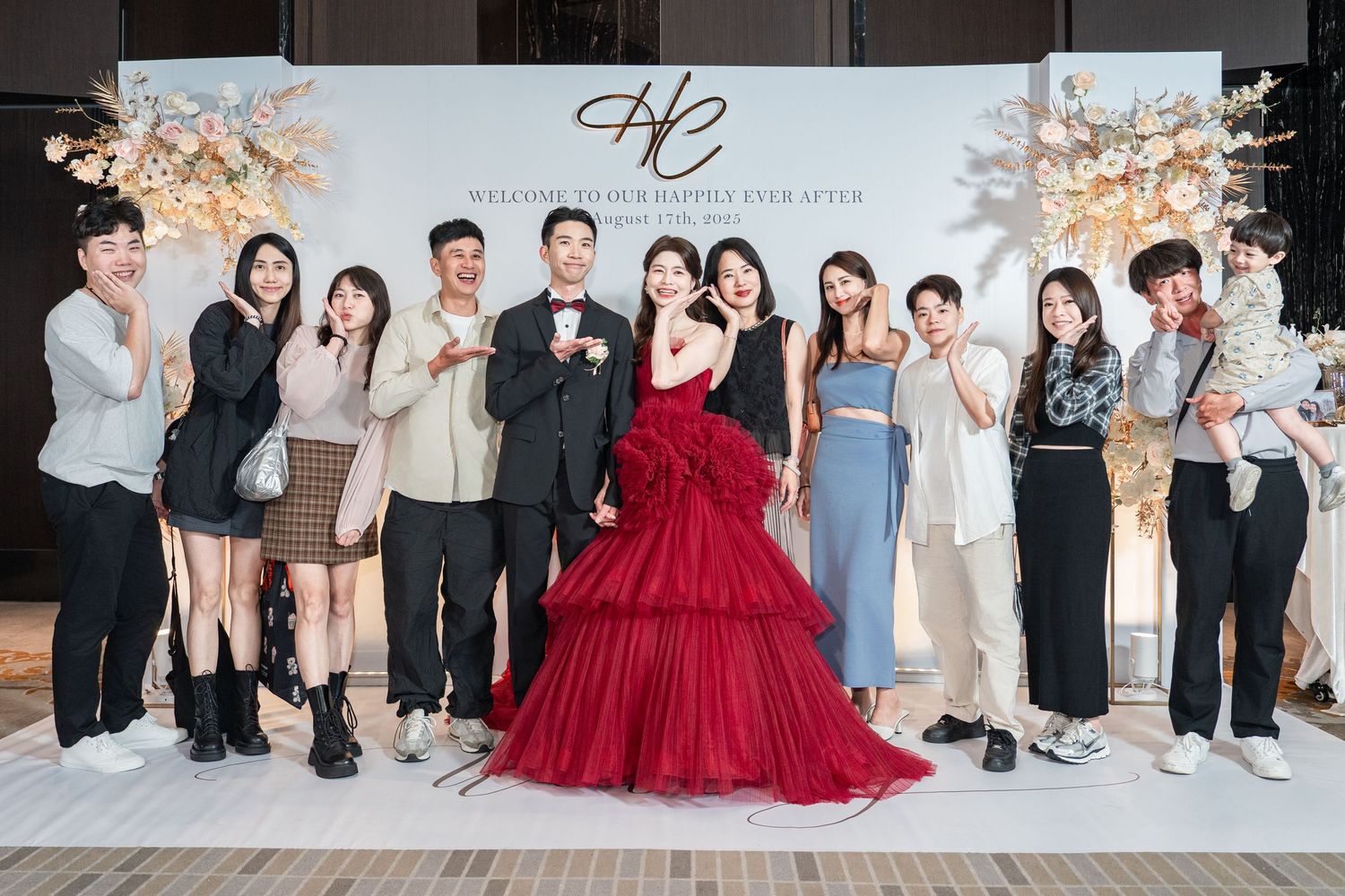 Wedding celebration group shot with people posing by decorative wall, featuring a stunning red tulle gown centerpiece.