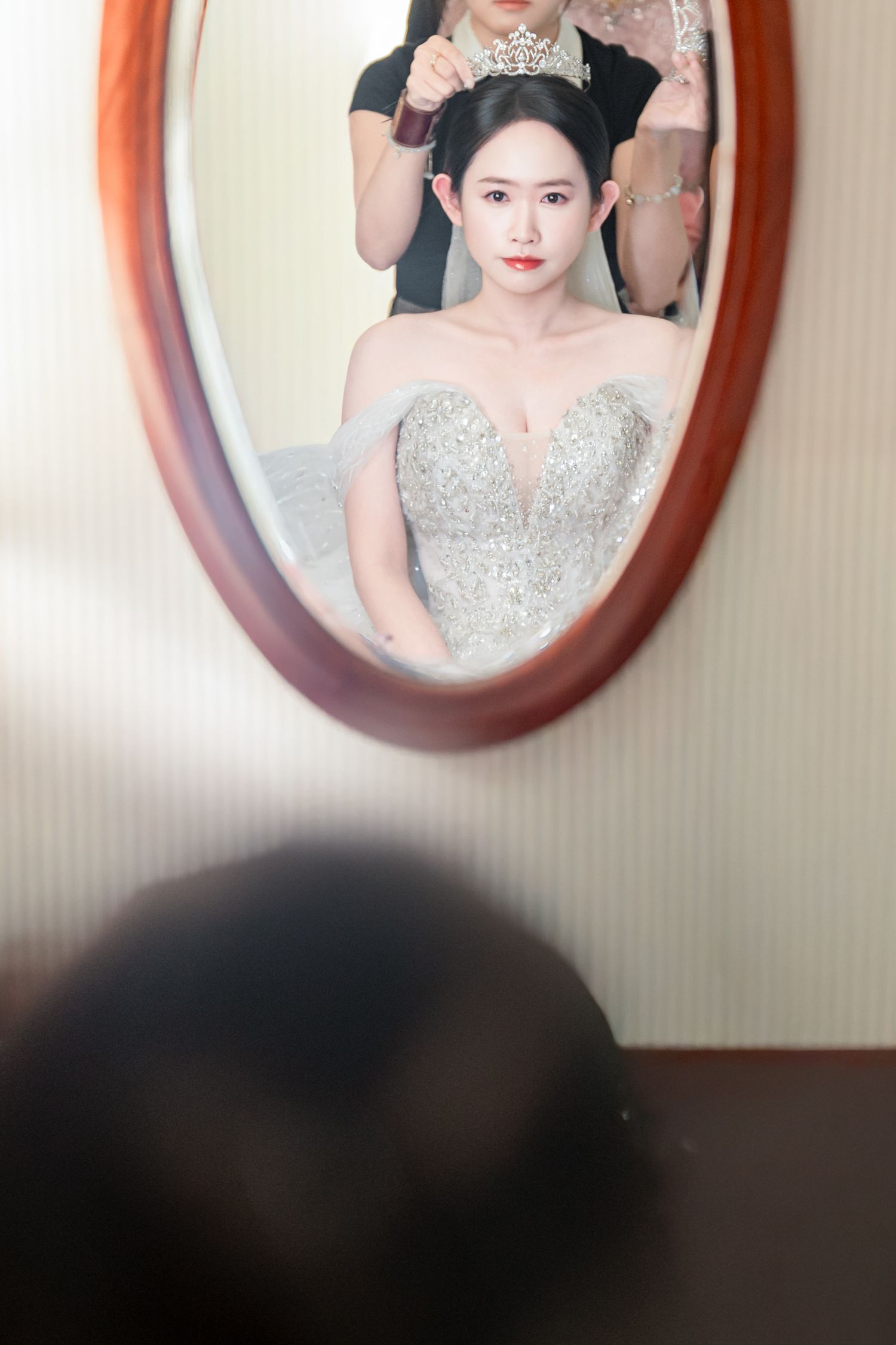 Mirror reflection shows a bride wearing a sparkly off-shoulder wedding gown while getting her hair styled.