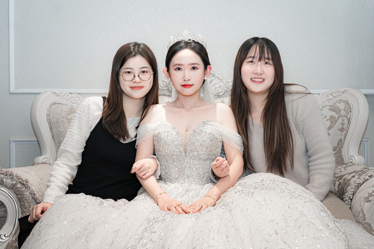 Three women in elegant white dresses pose together on a luxurious sofa in a studio setting.