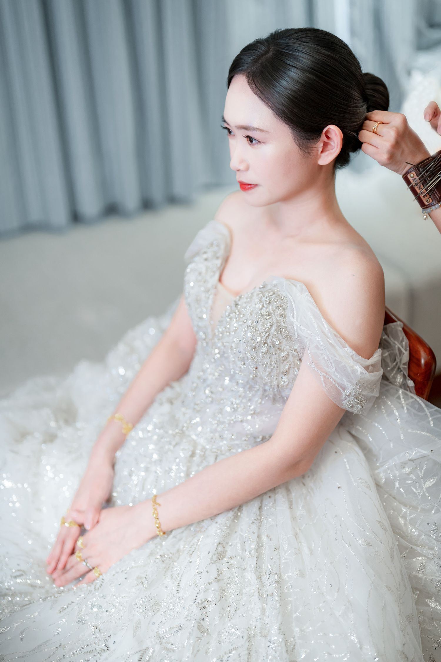 A bride in an elegant off-shoulder white wedding gown with sparkly details sits while getting ready for her special day.