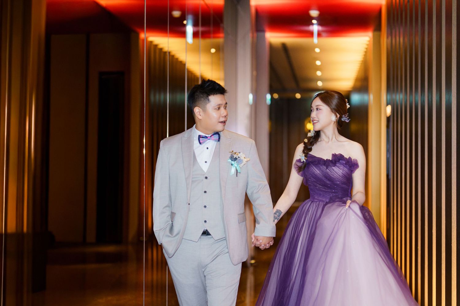 Couple in elegant evening attire walking through a warmly lit hotel hallway.