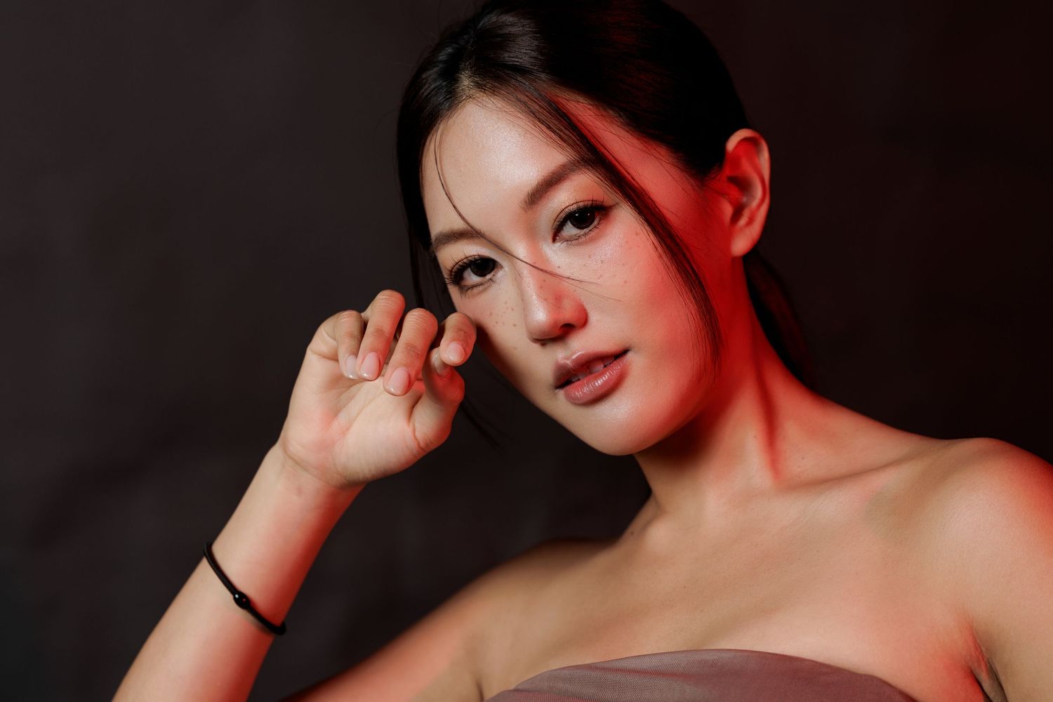 Young Asian woman with elegant makeup poses gracefully against a dark background with red lighting.