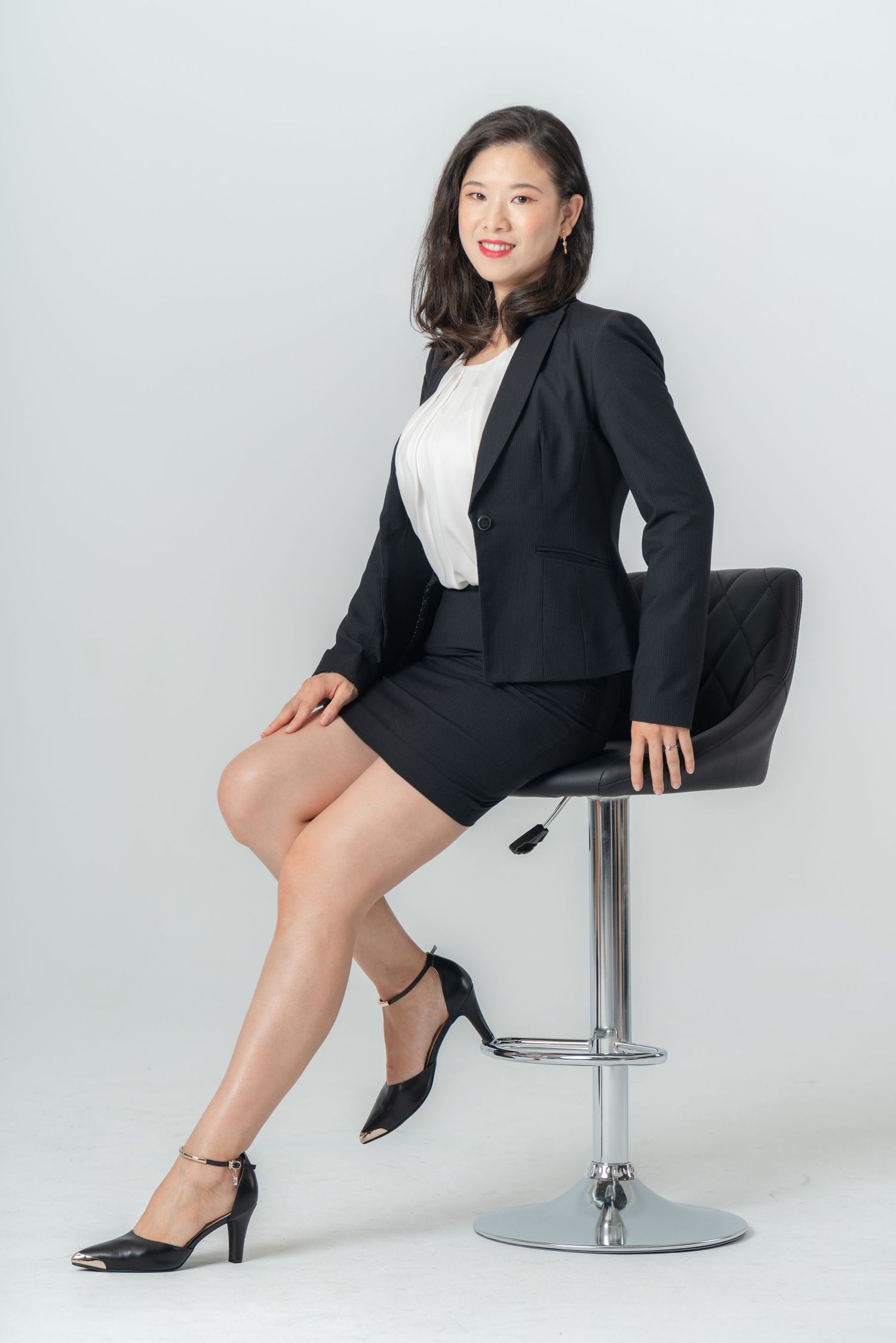Professional model in business attire poses on modern chrome bar stool against white studio background.