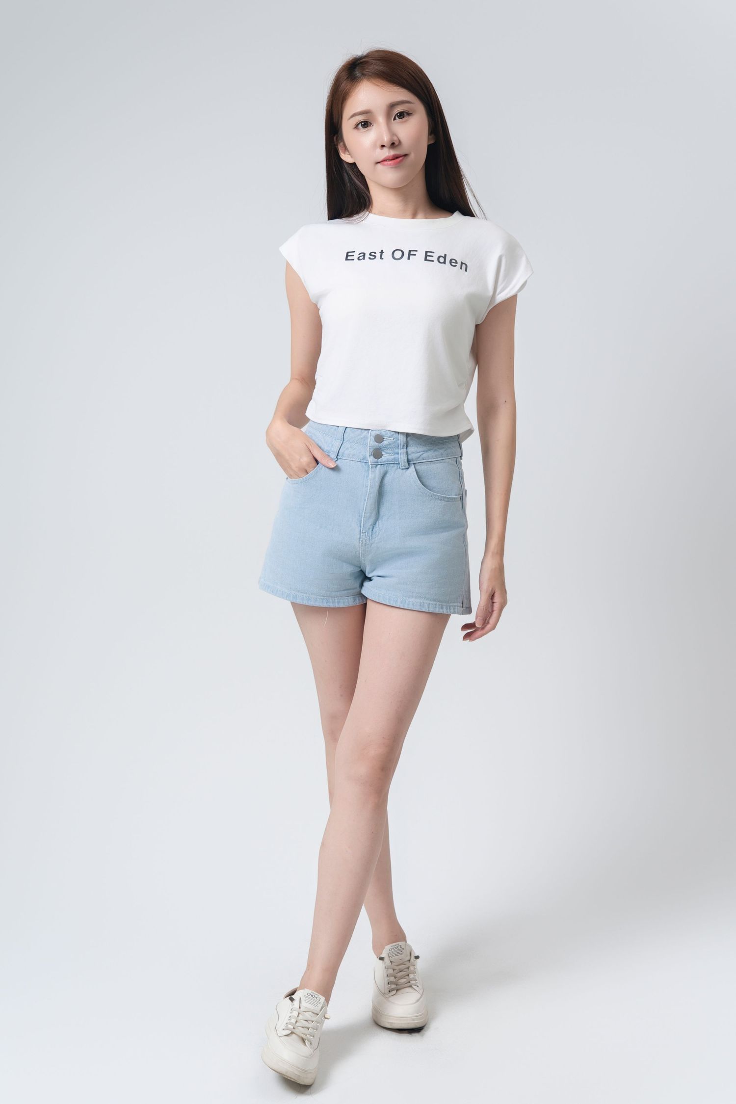 Model wearing light blue denim shorts and white graphic t-shirt with cream colored sneakers against white background.