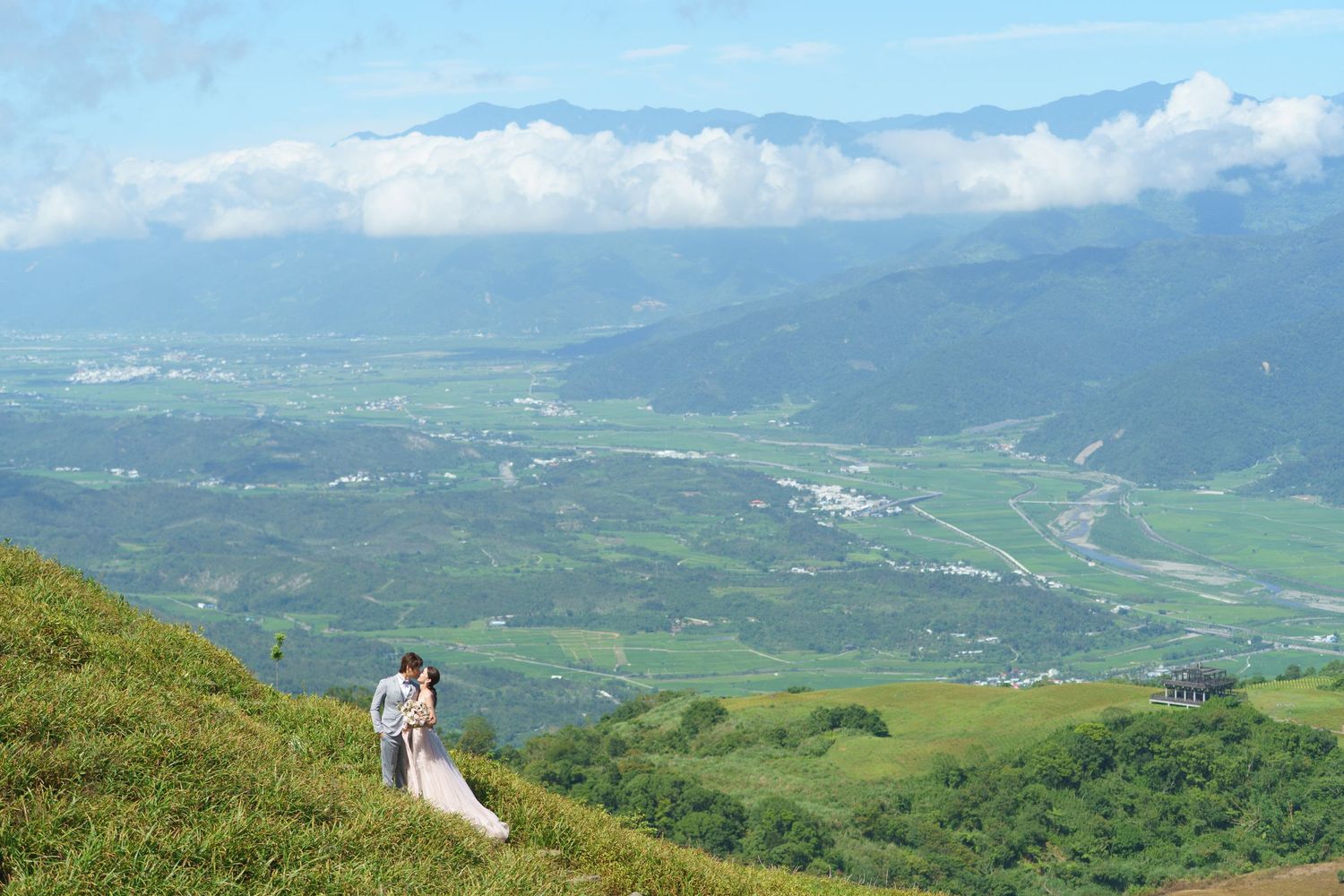 Panoramic mountain vista showing rolling green hills and valleys stretching towards distant snow-capped peaks on a sunny day.