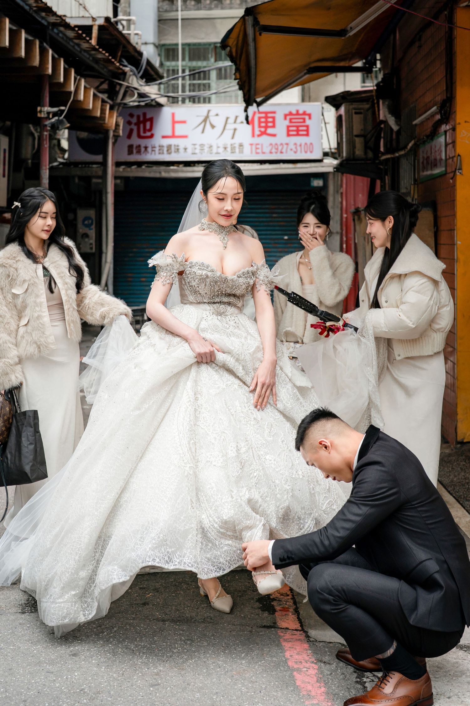 A wedding party in white dresses and suits poses playfully on a city street with Asian storefronts in the background.