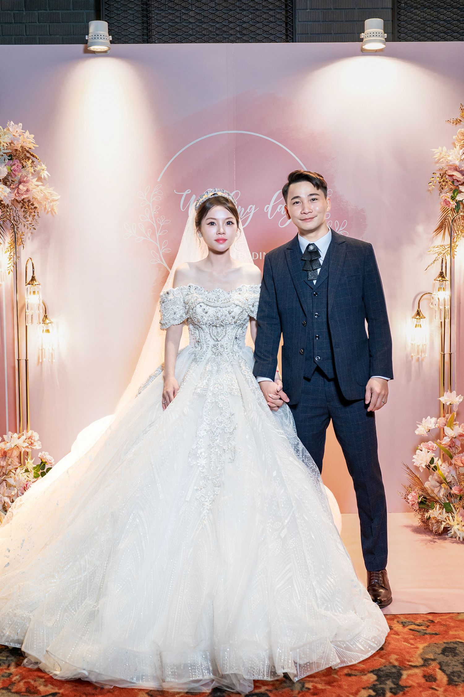 A wedding couple poses in an elegant venue with pink floral decor and white gowns against a romantic backdrop.