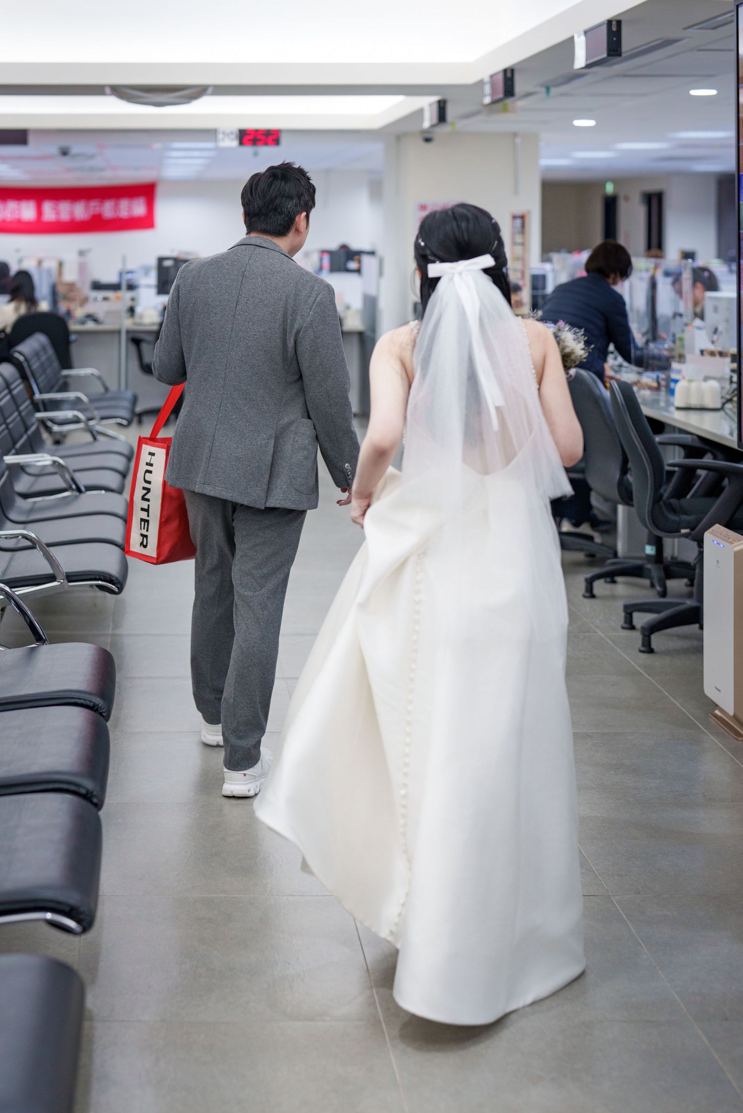 A couple in wedding attire walks through a modern office space with gray chairs and bright lighting.