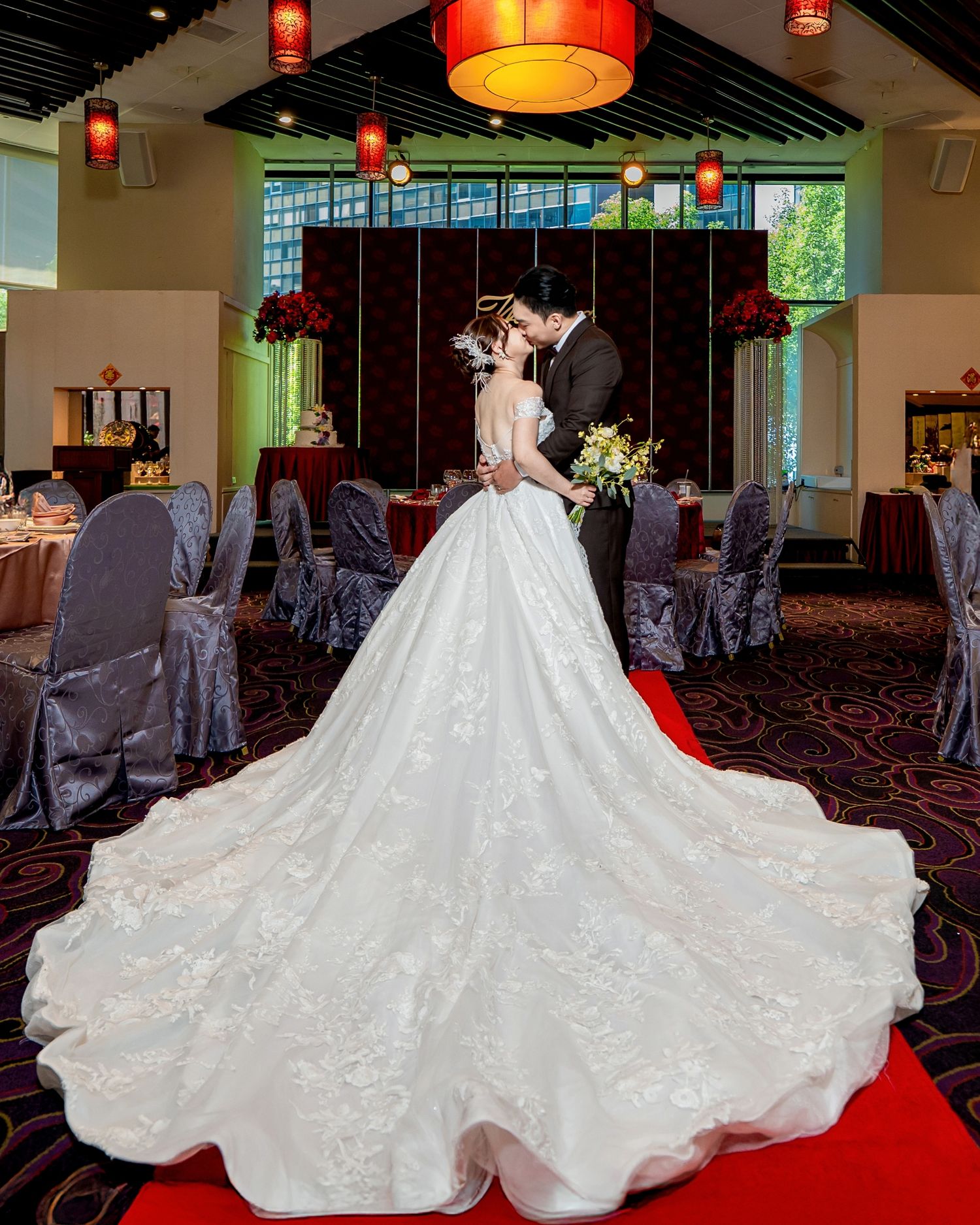 A wedding dress with a flowing white train is displayed in a banquet hall with red lighting and gray chair covers.