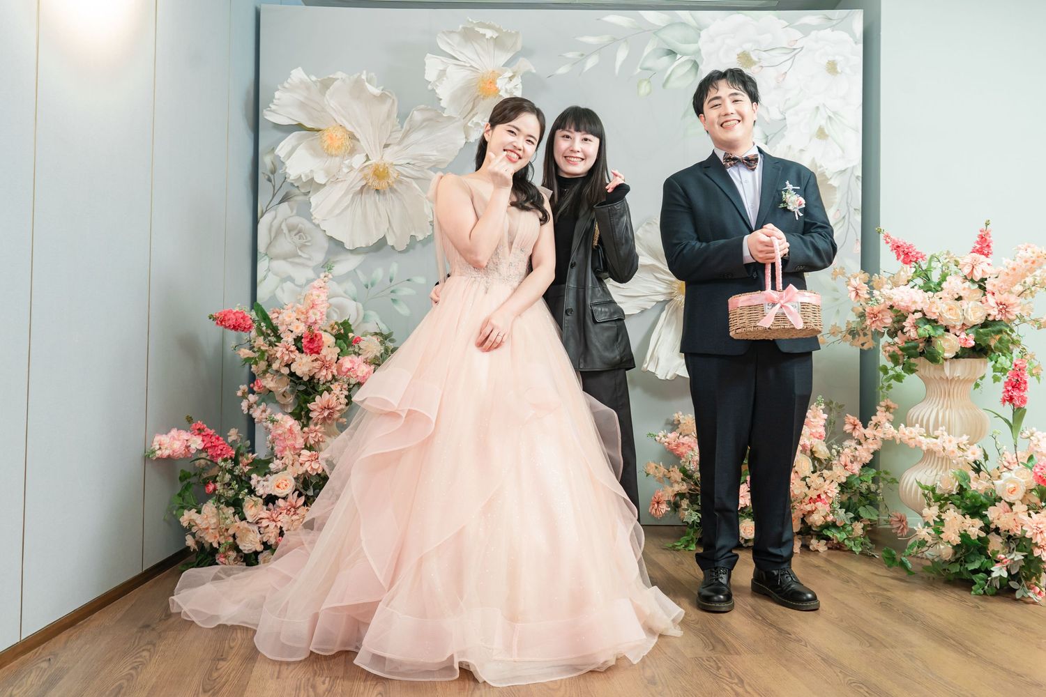 A series of wedding photos showing people in formal attire posing against a light blue wall with pink floral decorations.