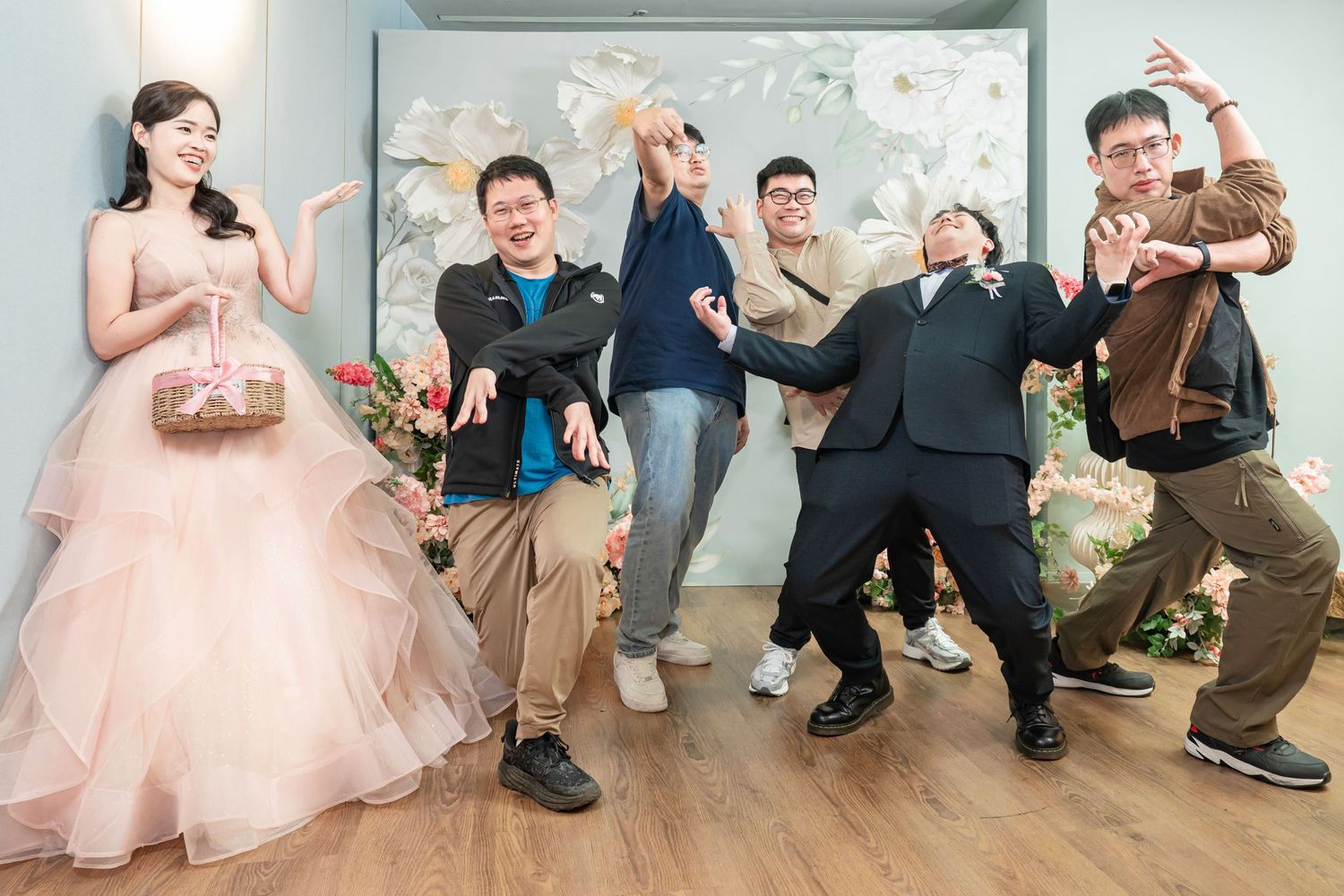 A group in casual and formal attire pose playfully in a studio setting with a pink tulle gown and decorative backdrop.