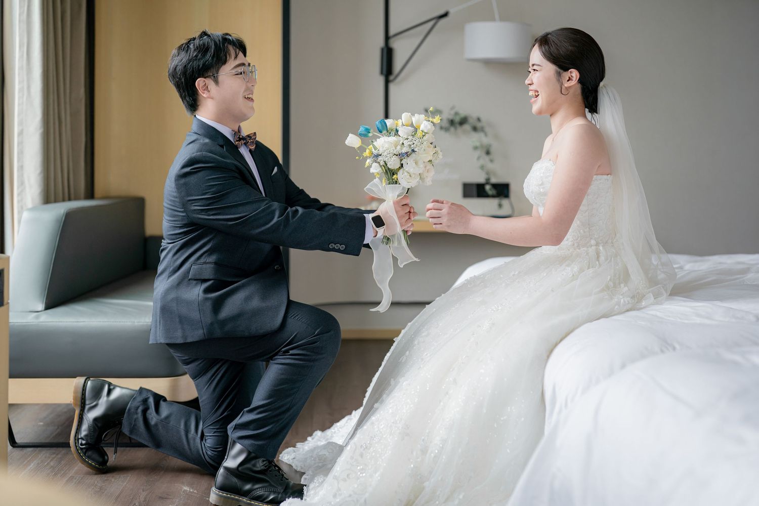 A couple in wedding attire share a tender moment as the groom kneels and presents a bouquet of white flowers to the bride.