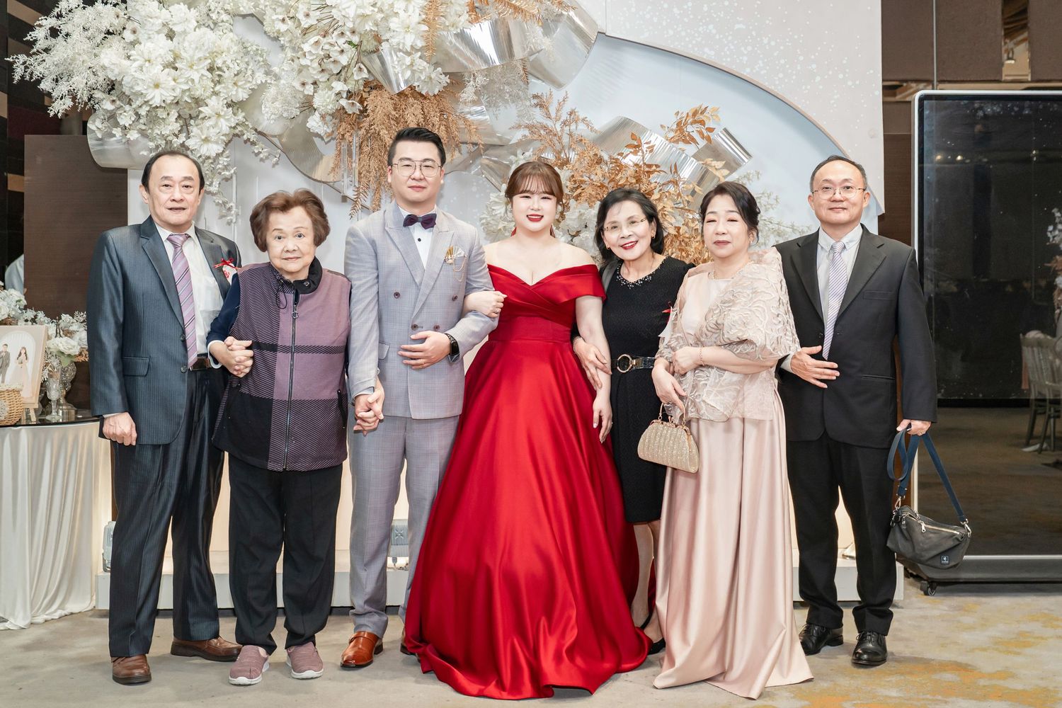Wedding party poses together in formal attire with a bride in red gown at an elegant indoor venue.