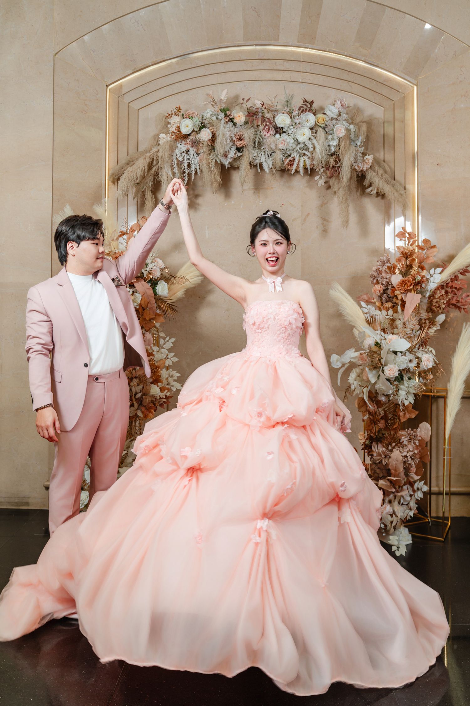 A couple celebrates in front of a floral arch with the person in a flowing peach ballgown gown raising their hands in joy.