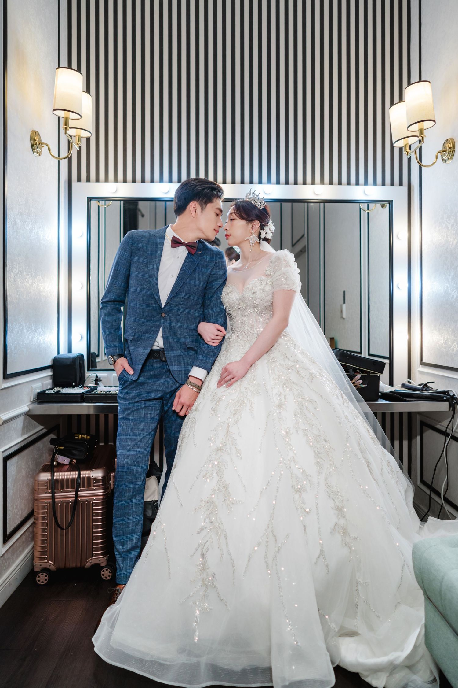 Elegant couple in formal wedding attire share a tender moment in a room with striped wallpaper and sconce lighting.