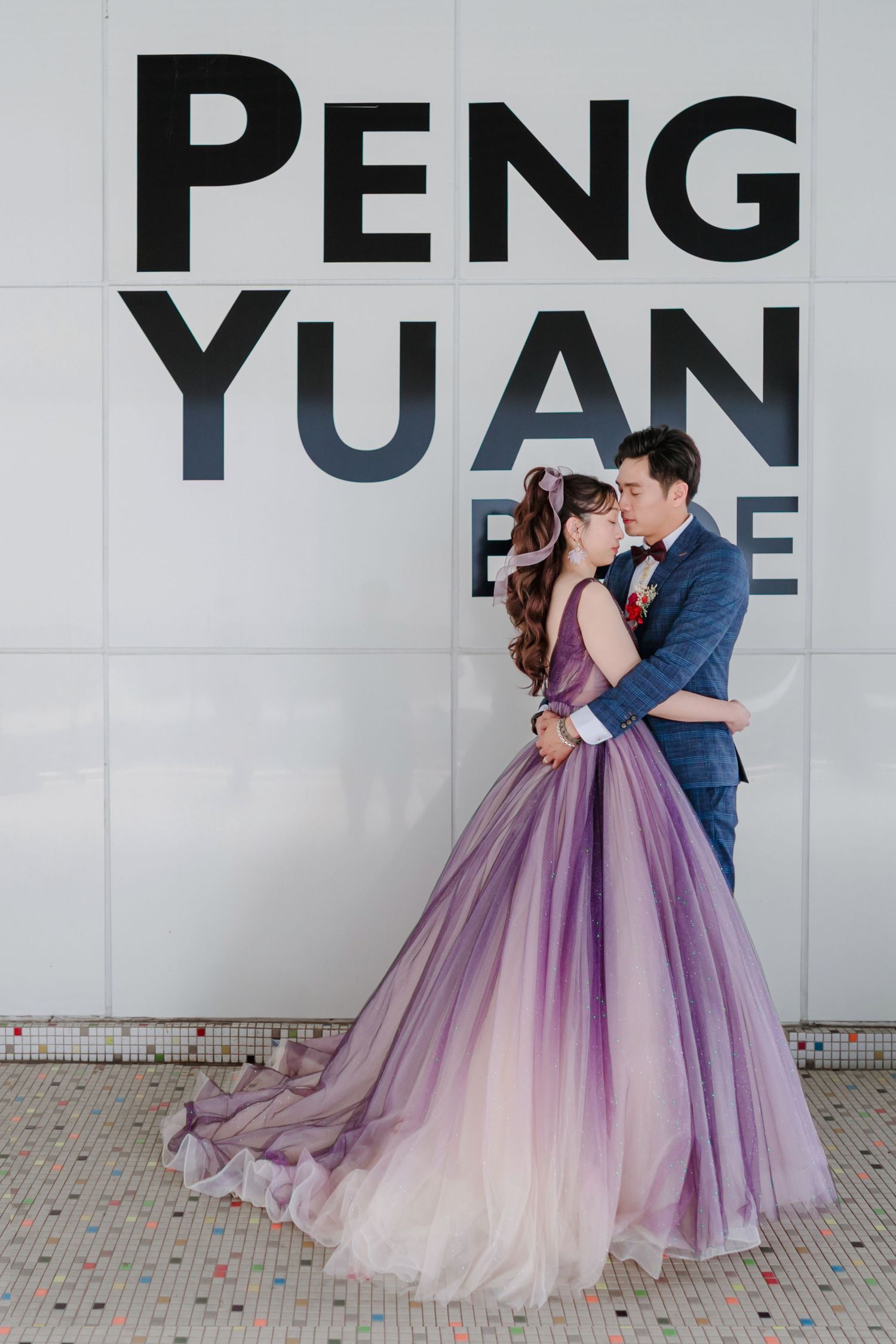 A couple in formal attire embrace against a wall with 'PENG YUAN' text, bride wearing a flowing purple ombre gown.