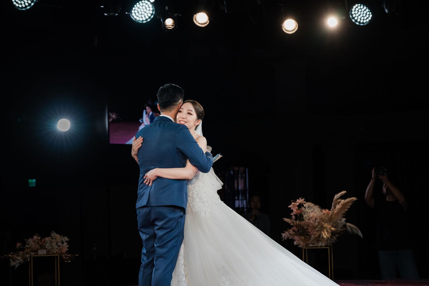 A couple shares their first dance as newlyweds on an illuminated stage with pampas grass decorations and twinkling lights.