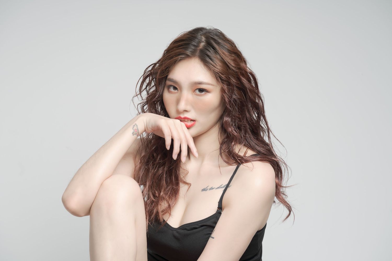 Studio portrait with soft lighting shows a person in black tank top with tousled brown hair and red lips posed against gray.