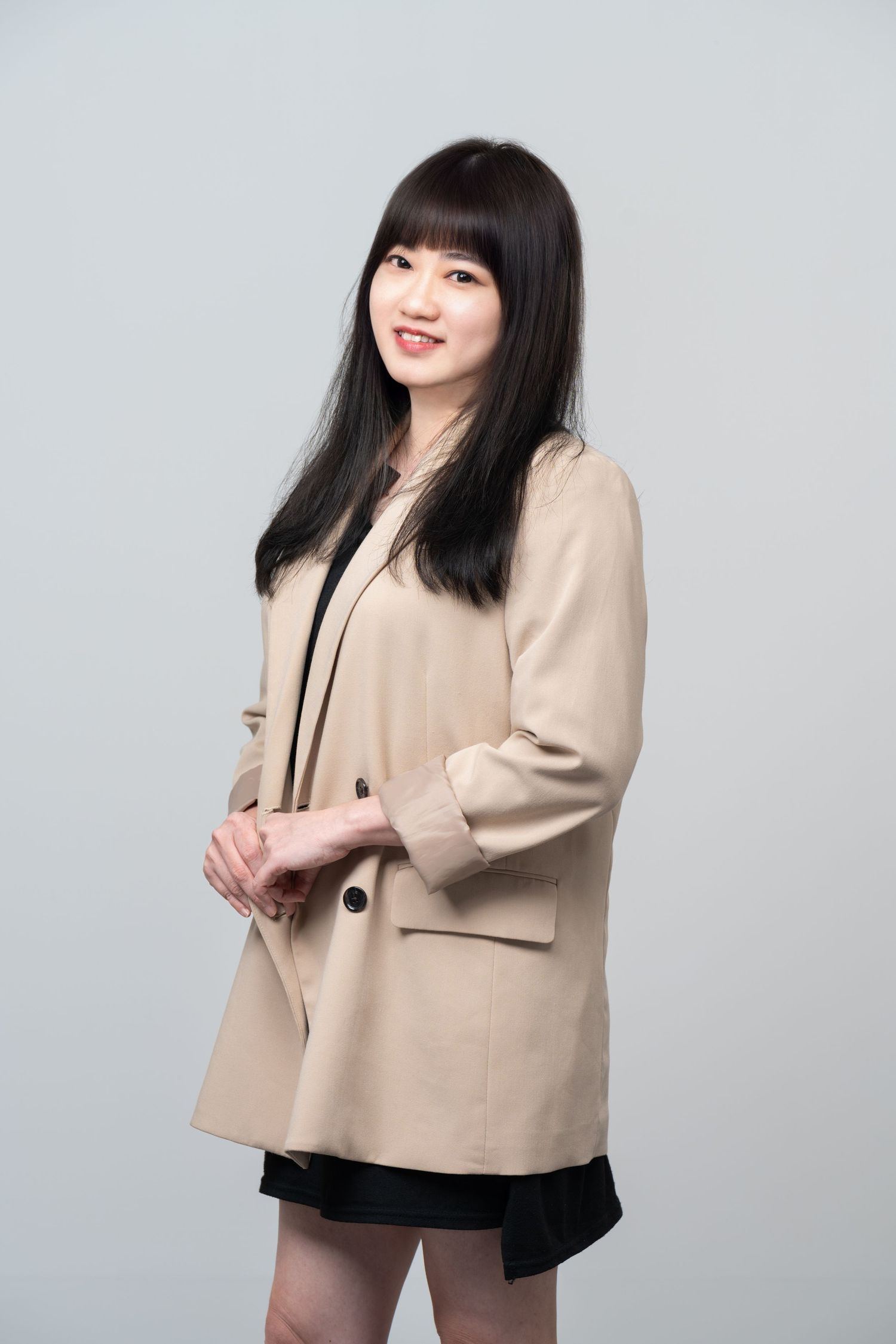A person wearing a beige blazer and black skirt poses against a gray background in a professional studio setting.