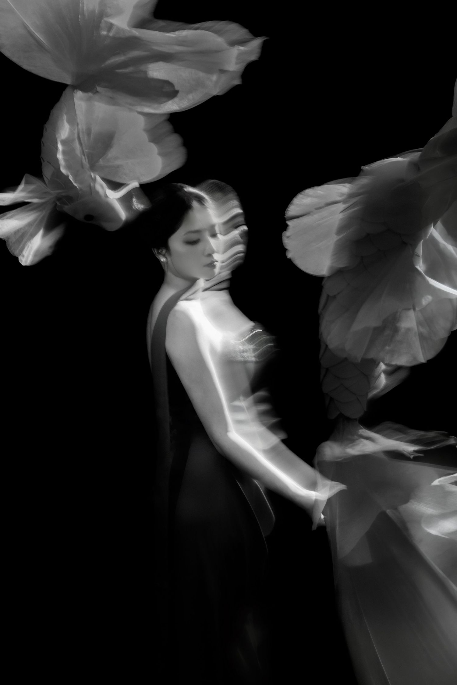 Black and white photo of a woman in an elegant gown surrounded by large flower petals against a dark background.
