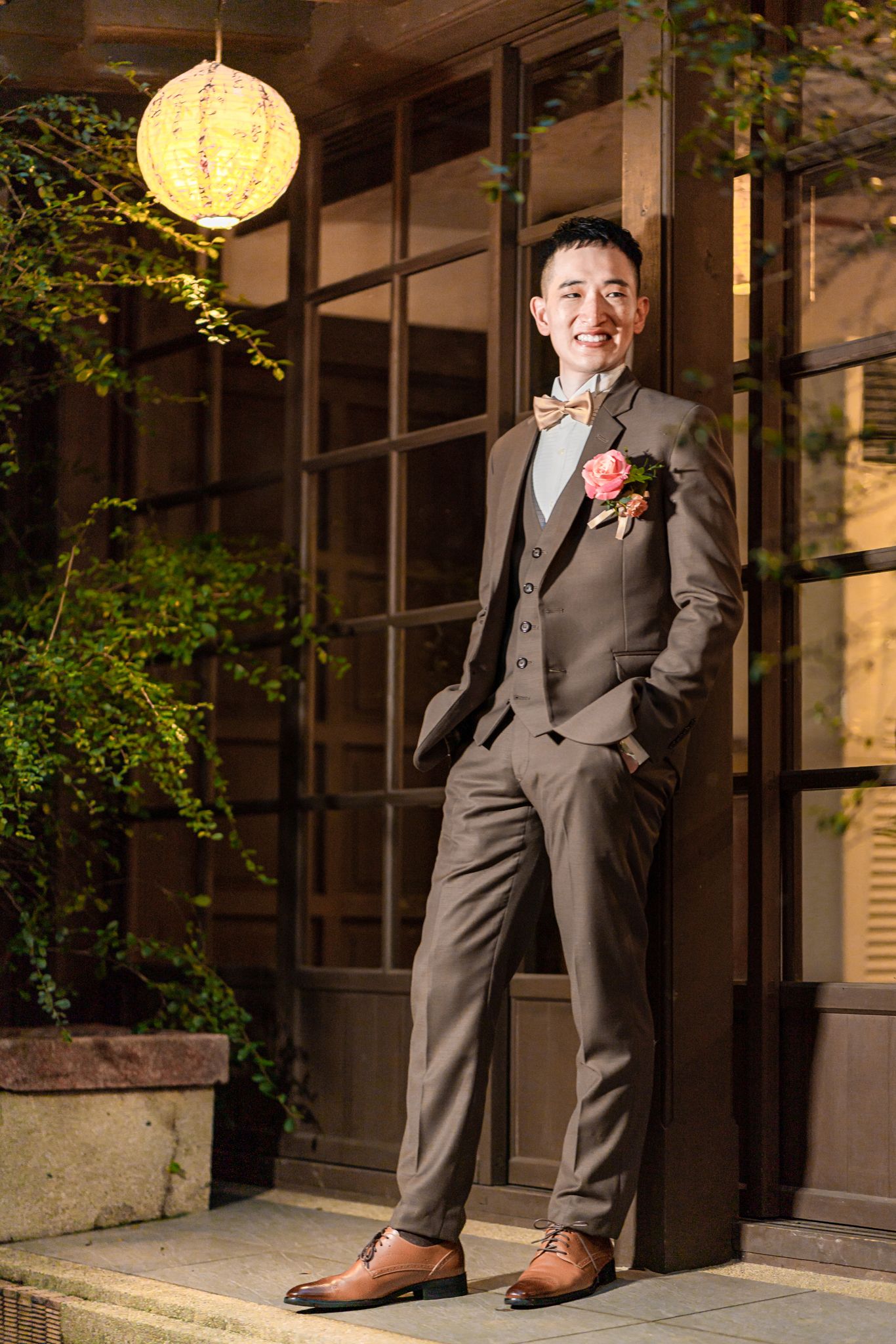 A groom in a brown suit and boutonniere poses confidently outside a building with a decorative lantern and window.